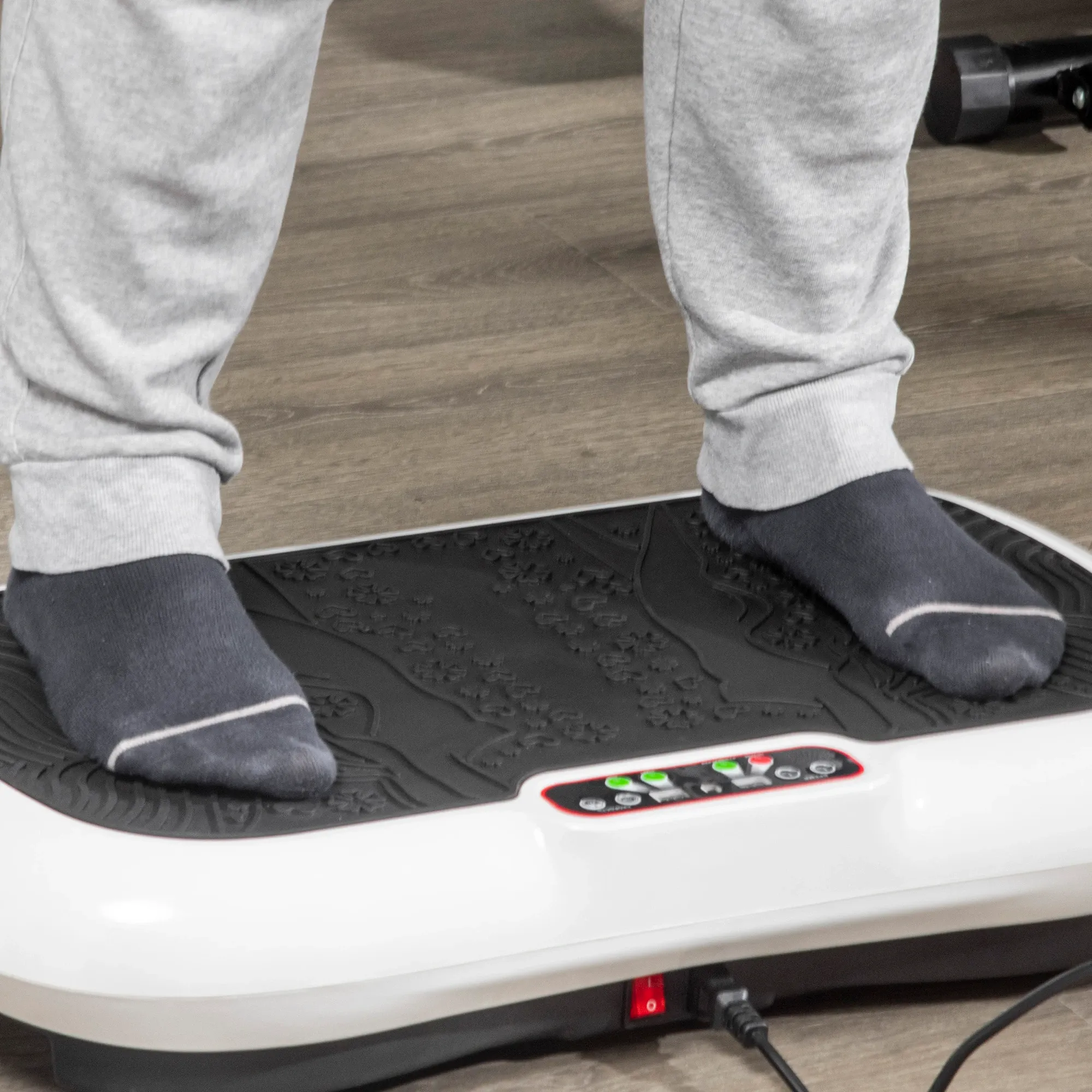 Beine und Füße einer Person auf einer vibrierenden Trainingsplattform. Graue Socken, Jogginghose. Schwarze Plattform mit weißer Basis. Holzfußboden.