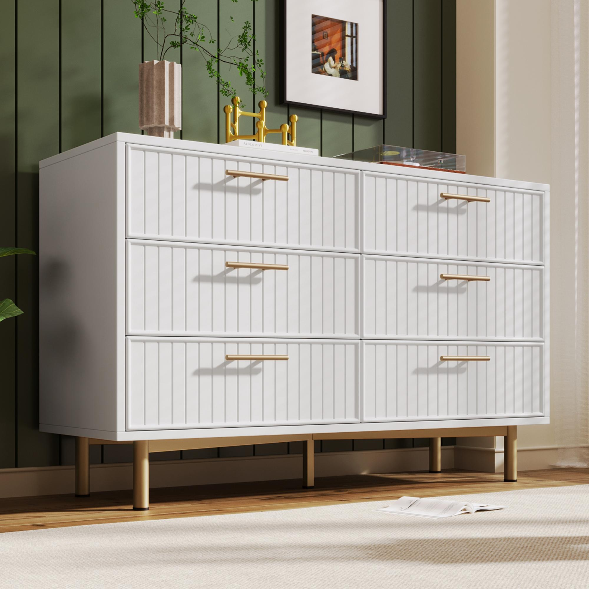 Weiße Kommode mit goldenen Griffen und Beinen. Grüner Wandhintergrund, gerahmtes Bild und Vase. Auf einem hellen Teppich.