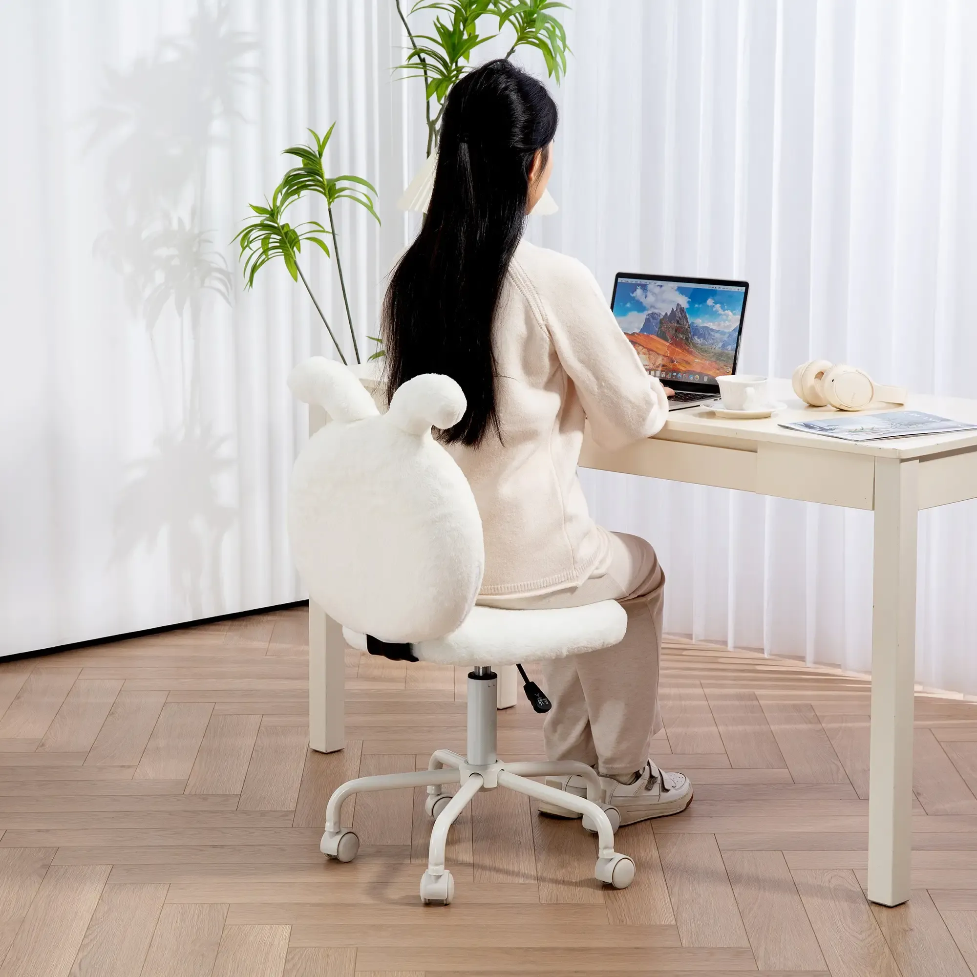 Eine Person sitzt an einem Schreibtisch und arbeitet an einem Laptop. Sie befindet sich in einem Raum mit weißen Vorhängen und einem weißen Stuhl.