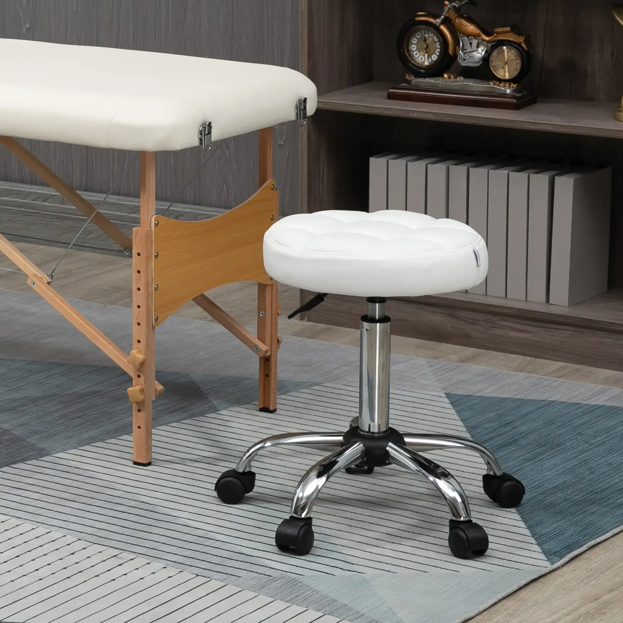 Eine Massageliege und ein weißer Hocker auf einem gemusterten Teppich. Ein Regal mit Büchern und einer Uhr im Hintergrund.