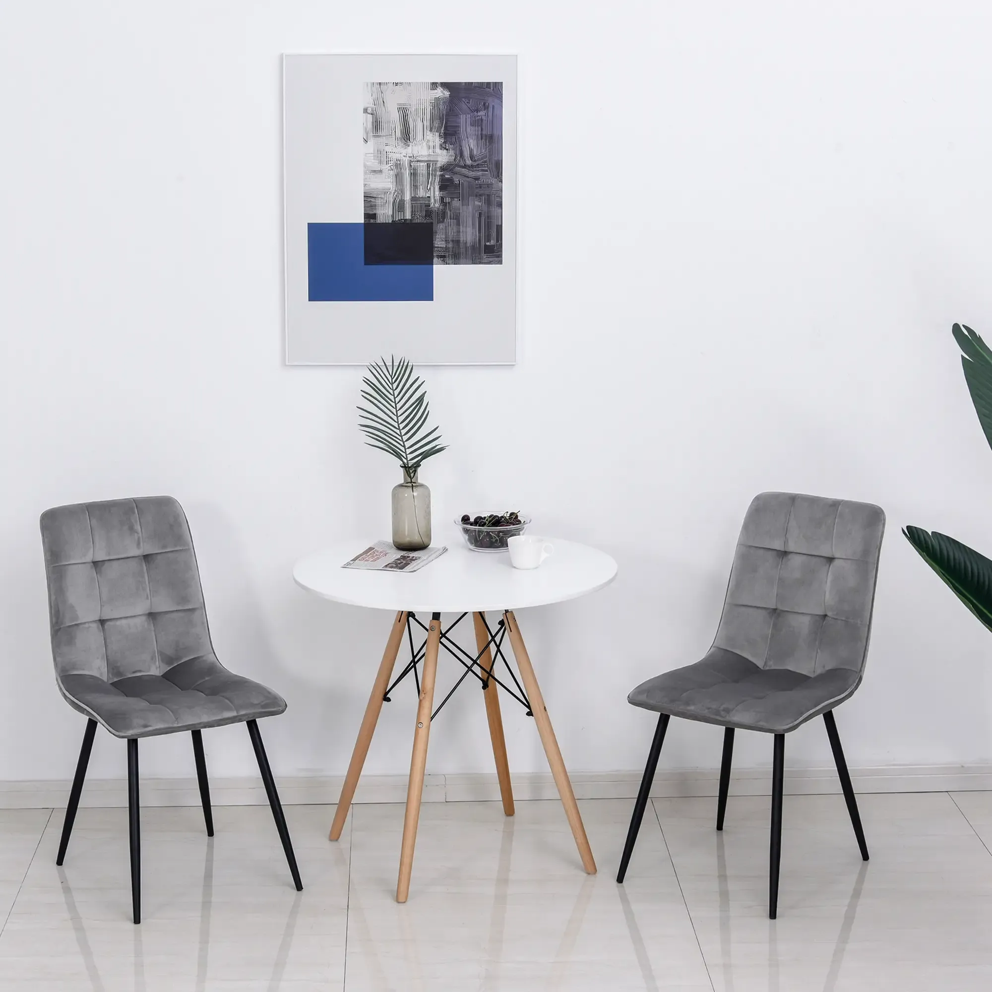 Zwei graue Stühle stehen einem weißen Tisch gegenüber. Ein Gemälde hängt an der weißen Wand. Eine Vase mit Blatt und eine Pflanze sind ebenfalls sichtbar.