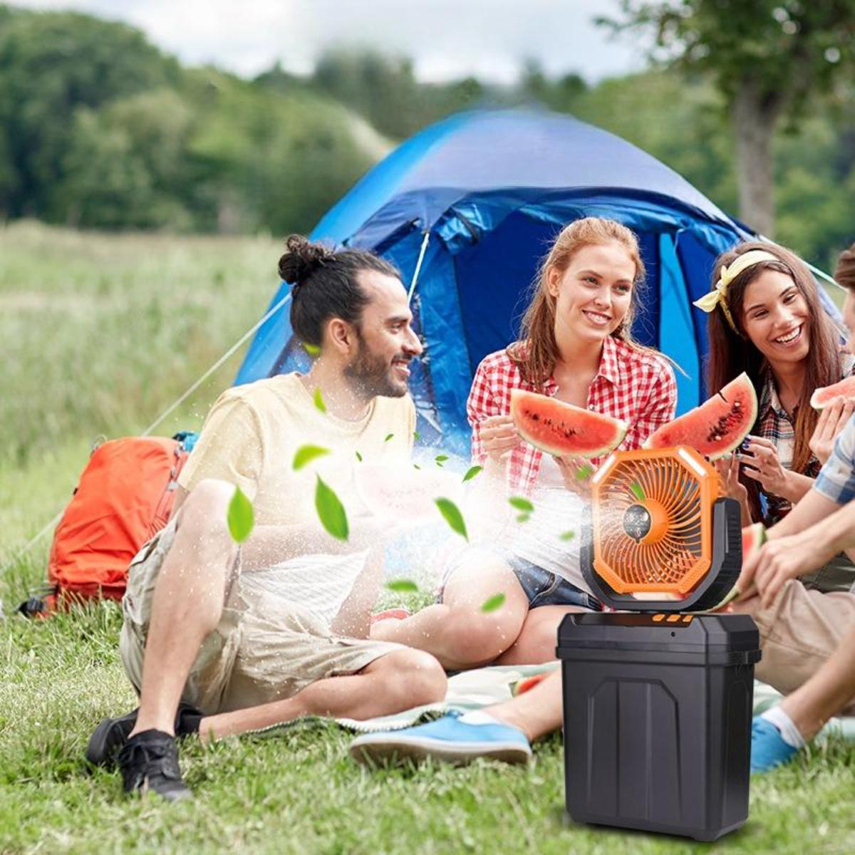 Eine Gruppe von Freunden auf einem Campingplatz mit einem Ventilator und Wassermelone. Menschen lächeln und genießen die Natur in der Nähe eines blauen Zeltes.