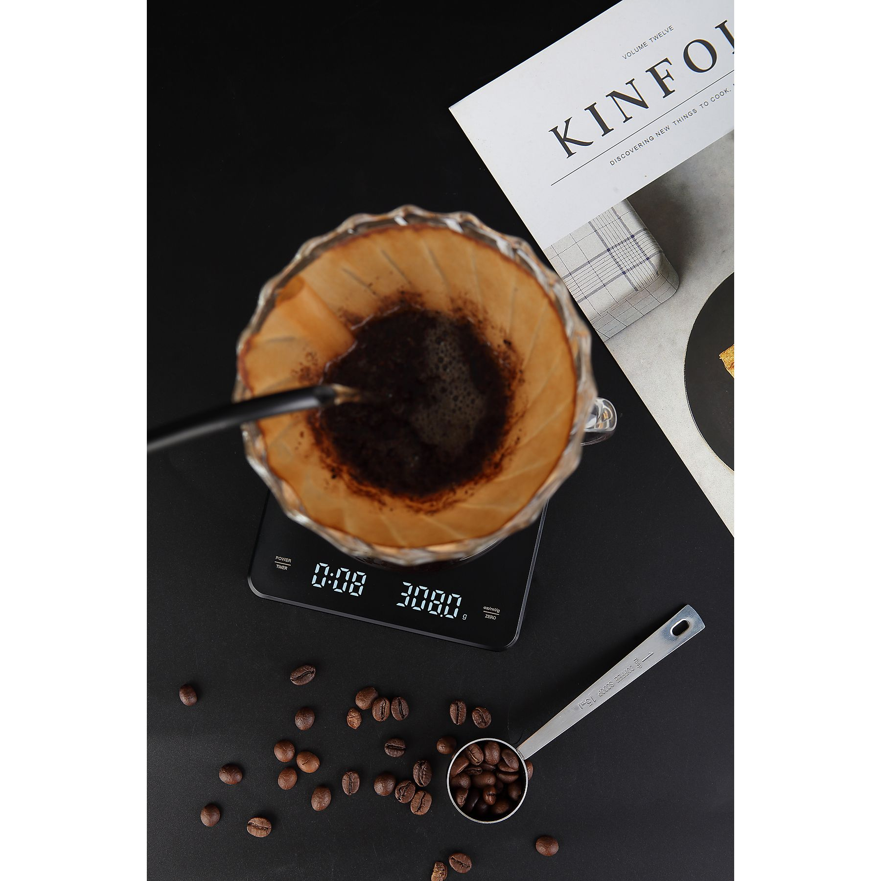 Kaffeezubereitung mit Glas-Pour-Over, digitaler Waage, Kaffeebohnen und einem Messlöffel auf schwarzer Oberfläche.