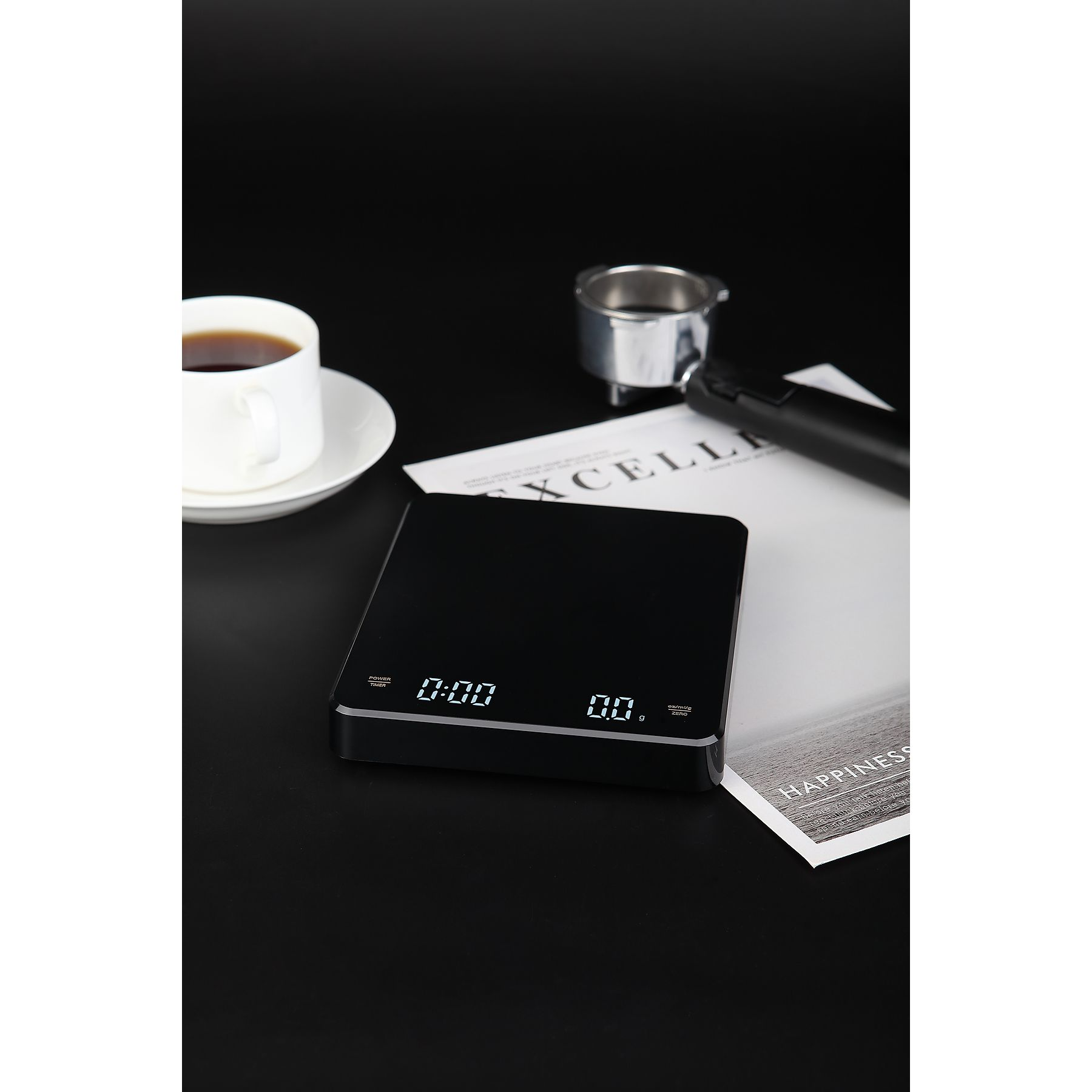 Schwarze Kaffeewaage mit Kaffeetasse, Zeitschrift und Siebträger auf schwarzer Oberfläche.