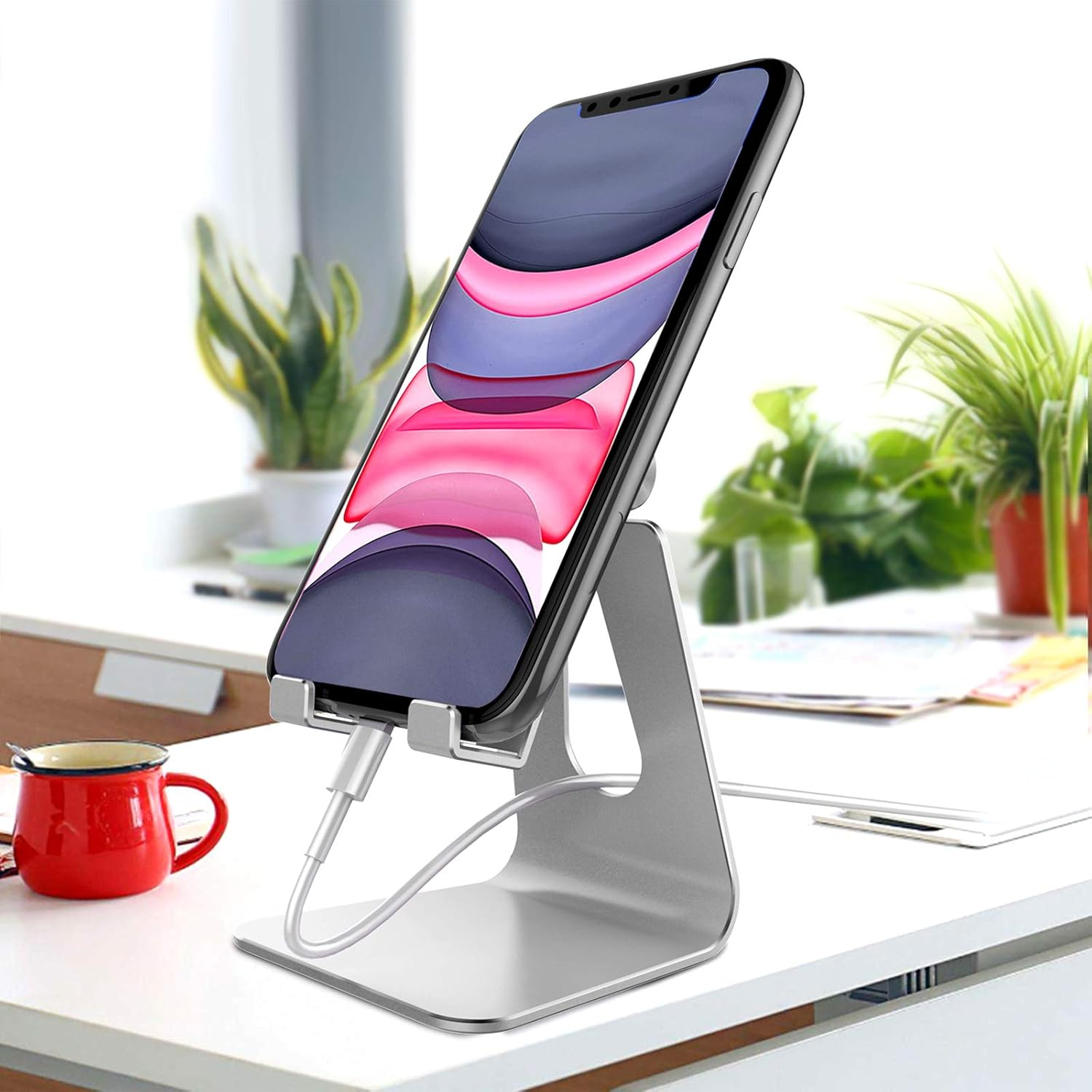 Ein Telefon auf einem silbernen Ständer. Eine rote Tasse, Pflanzen und ein weißer Schreibtisch füllen die Szene. Das Telefon zeigt bunte abstrakte Kunst.