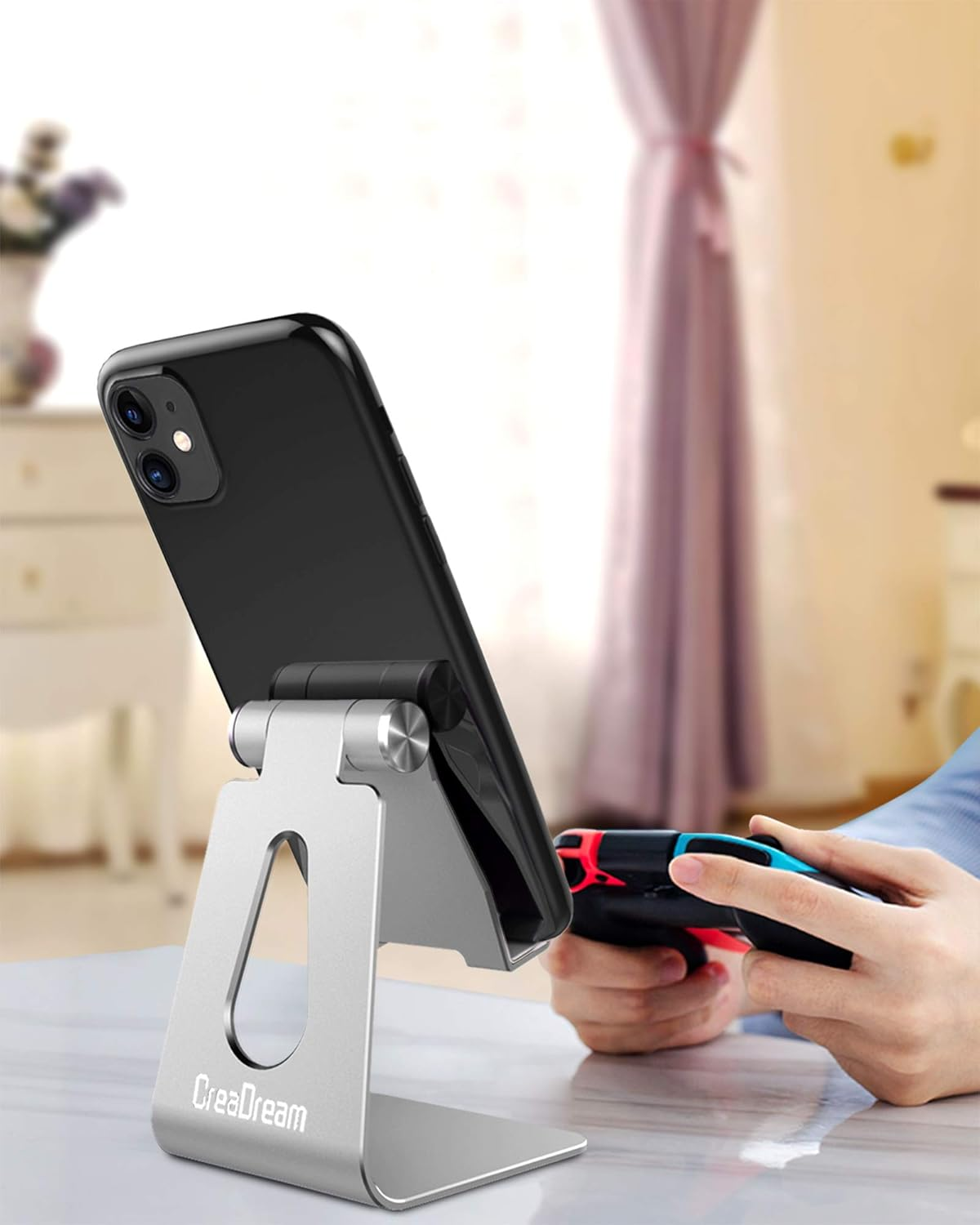 Ein schwarzes Telefon auf einem silbernen Ständer wird gehalten. Eine Person hält einen Gamecontroller. Der Hintergrund zeigt einen verschwommenen Raum.