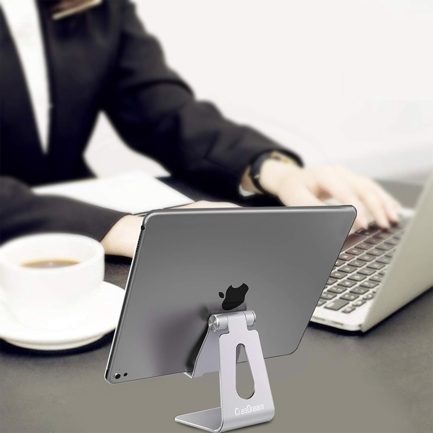 Ein Tablet in einem silbernen Ständer, eine Person, die an einem Laptop arbeitet, und eine Tasse Kaffee auf dem Tisch.