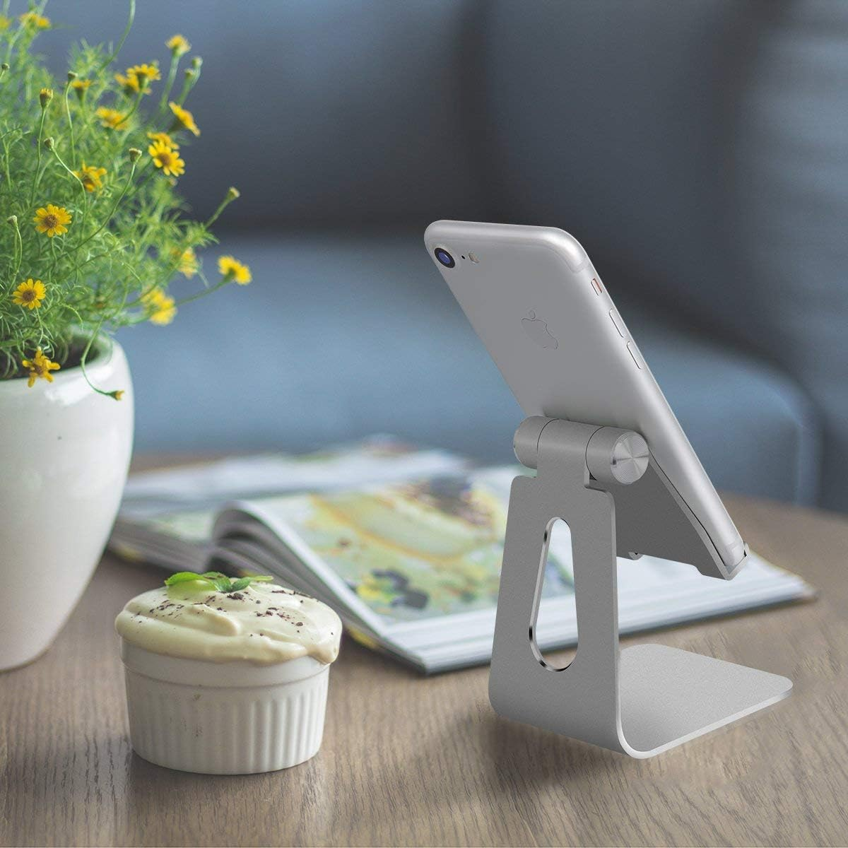 Silbernes Telefon auf Ständer, Dessert, Blumen und Zeitschrift auf einem Holztisch. Hintergrund ist eine verschwommene blaue Wand.