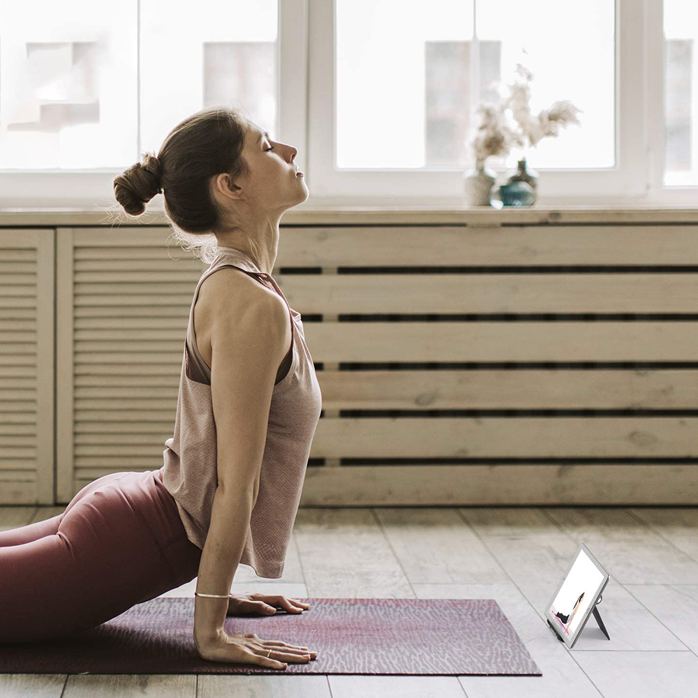 Frau in Yoga-Pose auf einer Matte, blickt mit geschlossenen Augen nach oben. Tablet zeigt Yoga-Session. Holzwand im Hintergrund.
