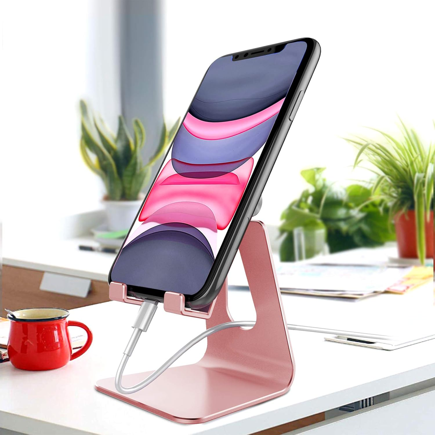 Ein Telefon steht auf einem rosa Ständer auf einem weißen Schreibtisch. Links ist eine rote Tasse, im Hintergrund Pflanzen.