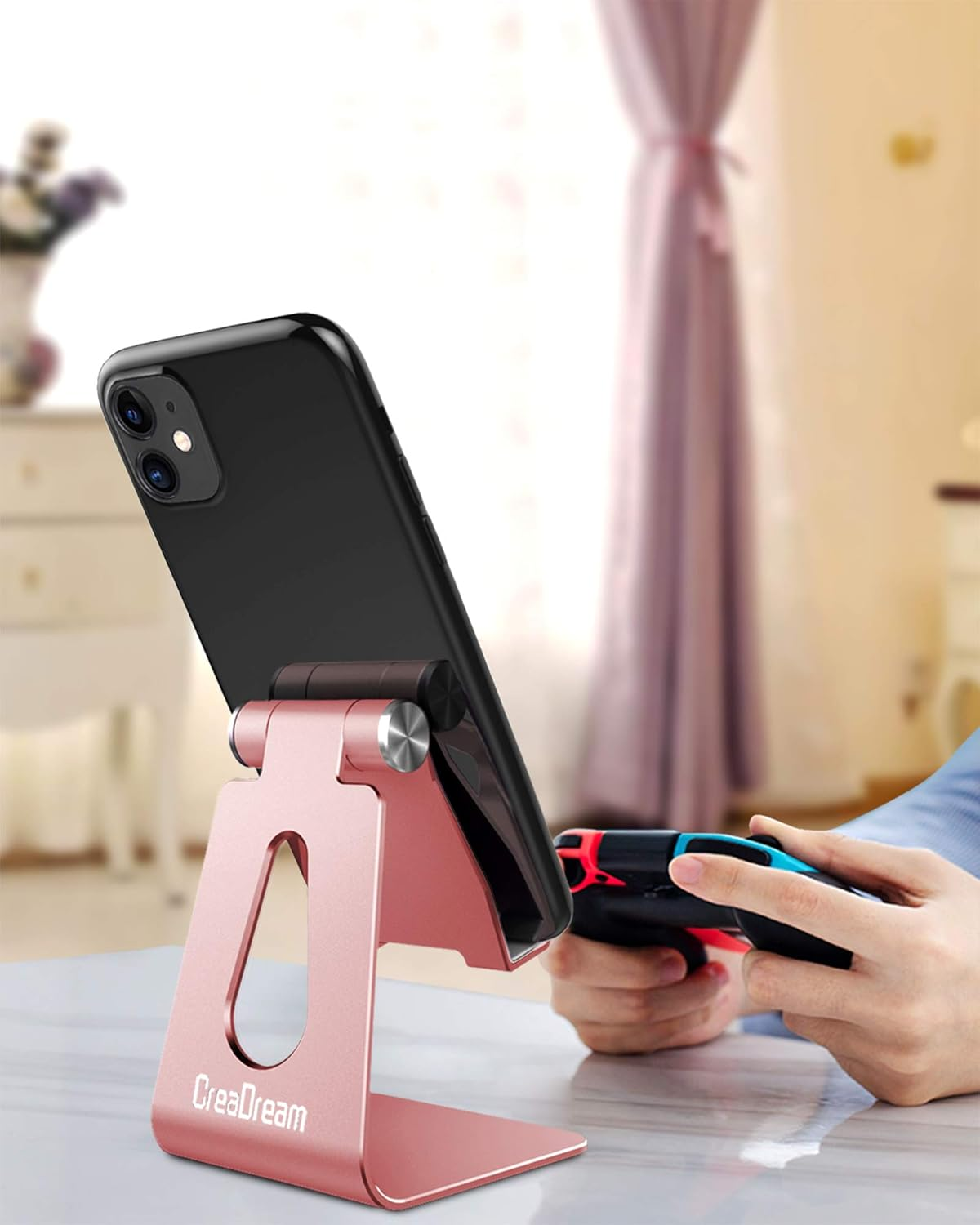 Ein schwarzes Smartphone steht auf einem roségoldenen Ständer. Eine Person hält einen Game-Controller. Im Hintergrund Vorhang und Tisch.