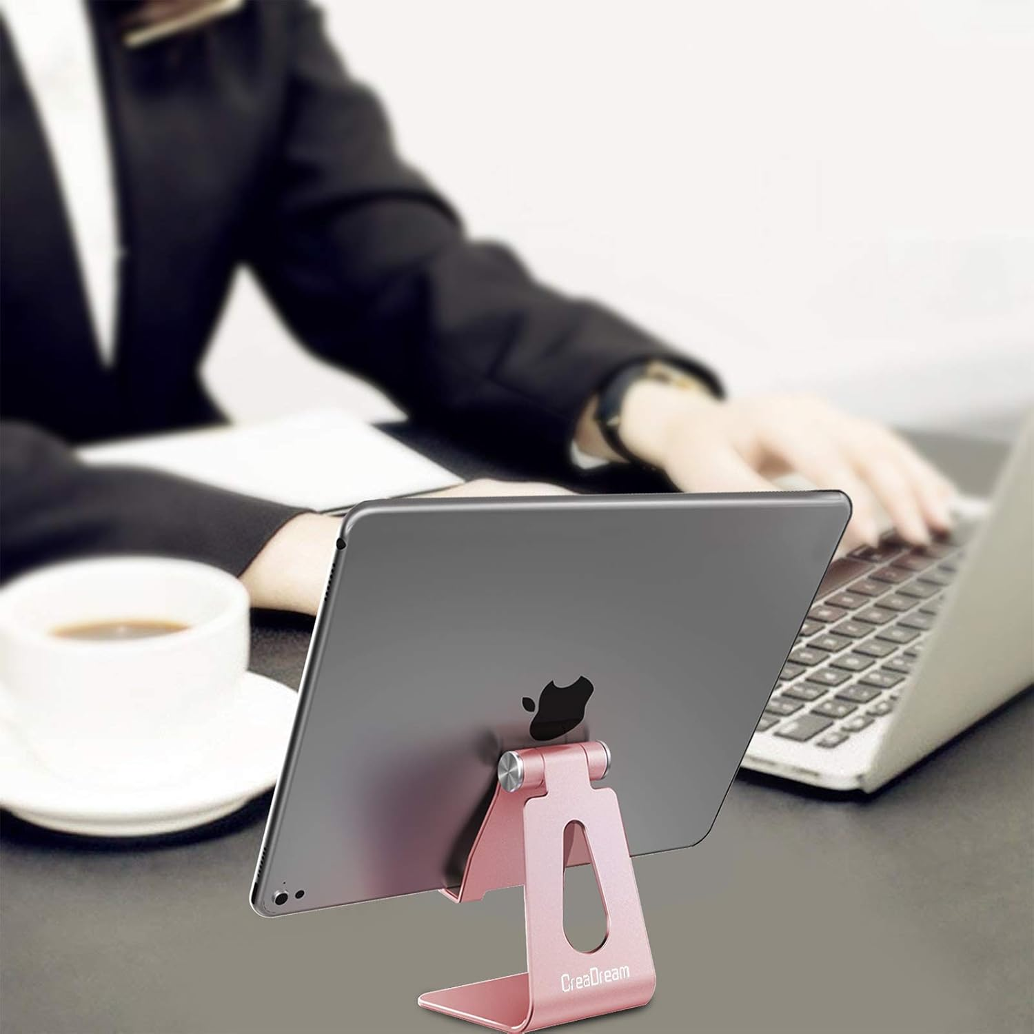 Ein roségoldener Tablet-Ständer hält ein Tablet. Im verschwommenen Hintergrund tippt eine Person auf einem Laptop. Links steht eine Kaffeetasse.