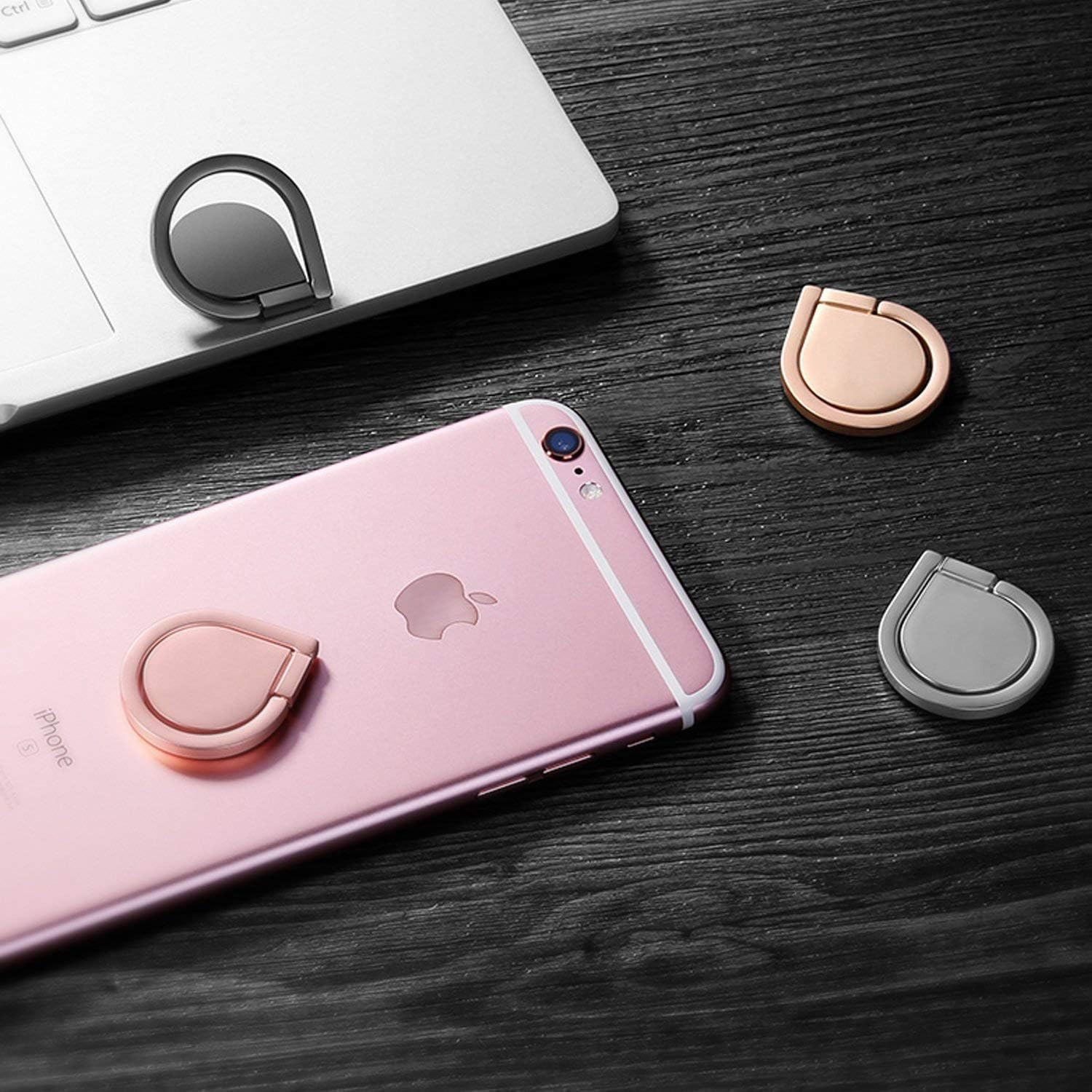 Ein pinkfarbenes iPhone mit einem roségoldenen Ringständer, drei Ringständern und einem Laptop liegen auf einer dunklen Holzoberfläche.