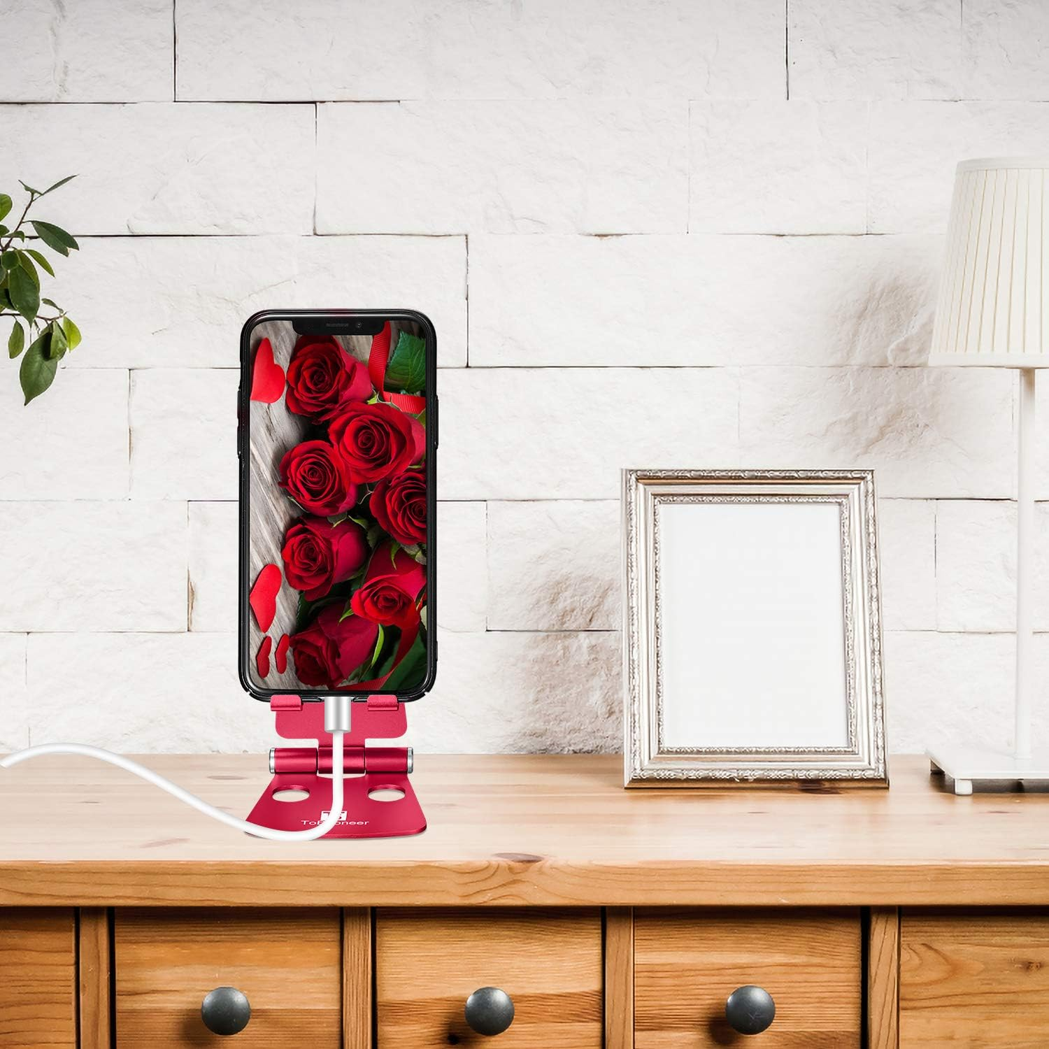 Ein Telefon mit Rosen in einem roten Ständer auf einem Holztisch. Ein weißes Kabel, eine Lampe und ein Rahmen sind ebenfalls vorhanden.