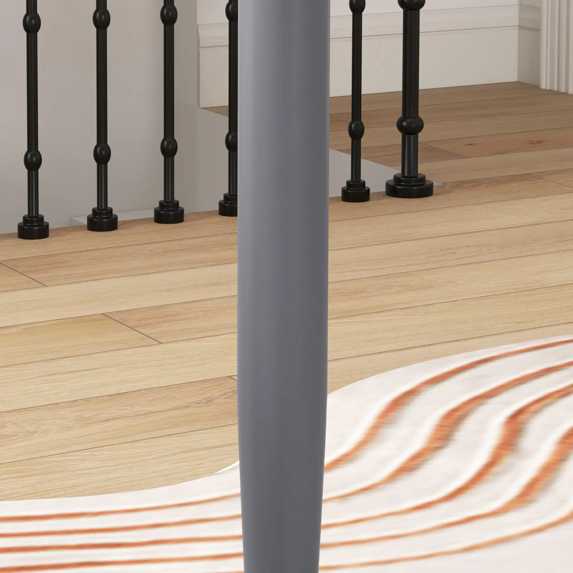 Eine graue, zylindrische Säule steht vor einem Holzboden und schwarzen Balustern. Darunter liegt ein Teppich mit orangefarbenen Streifen.