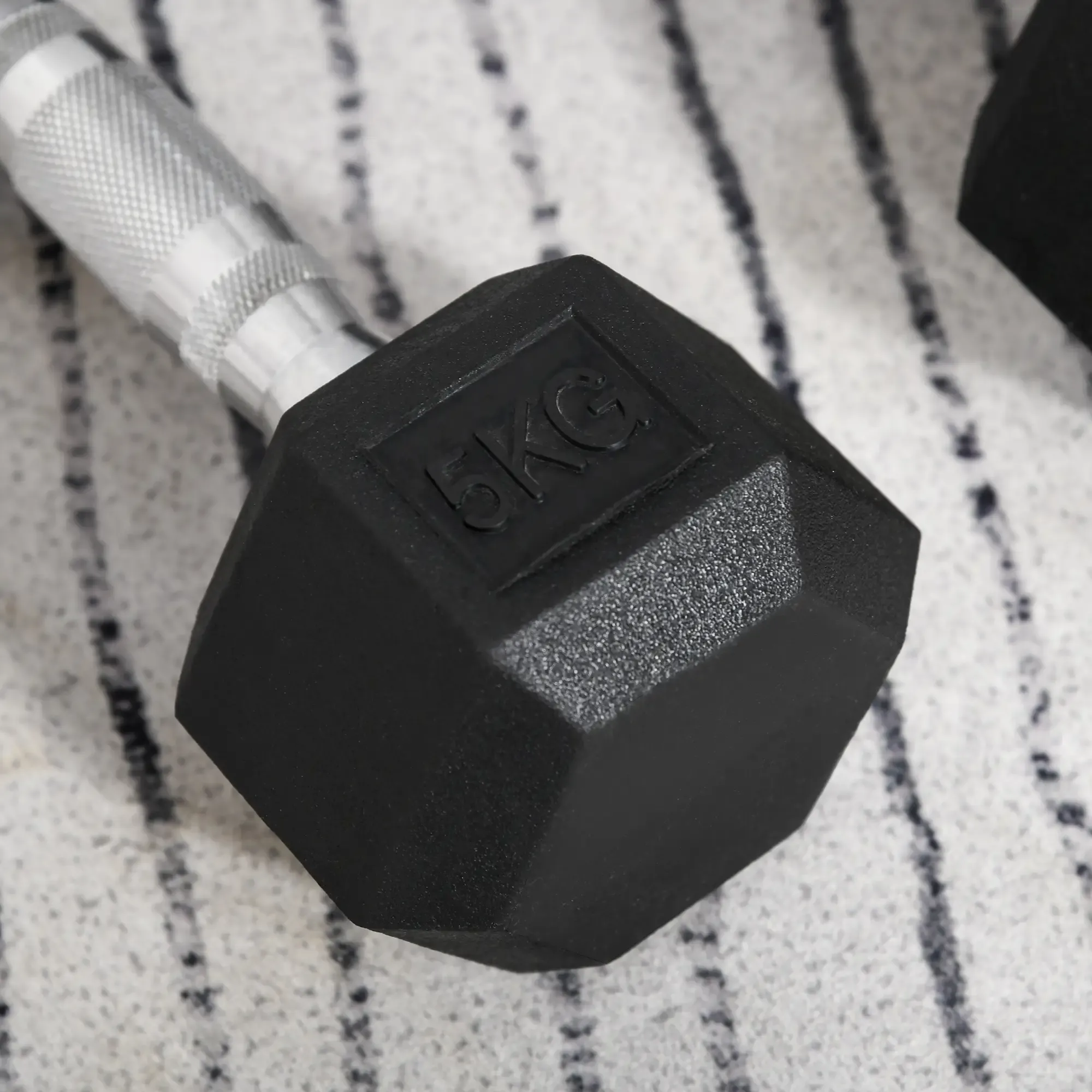 Eine schwarze 5-KG-Hantel liegt auf einer gemusterten Oberfläche. Der metallische Griff ist sichtbar. Das Gewicht ist aufgedruckt.