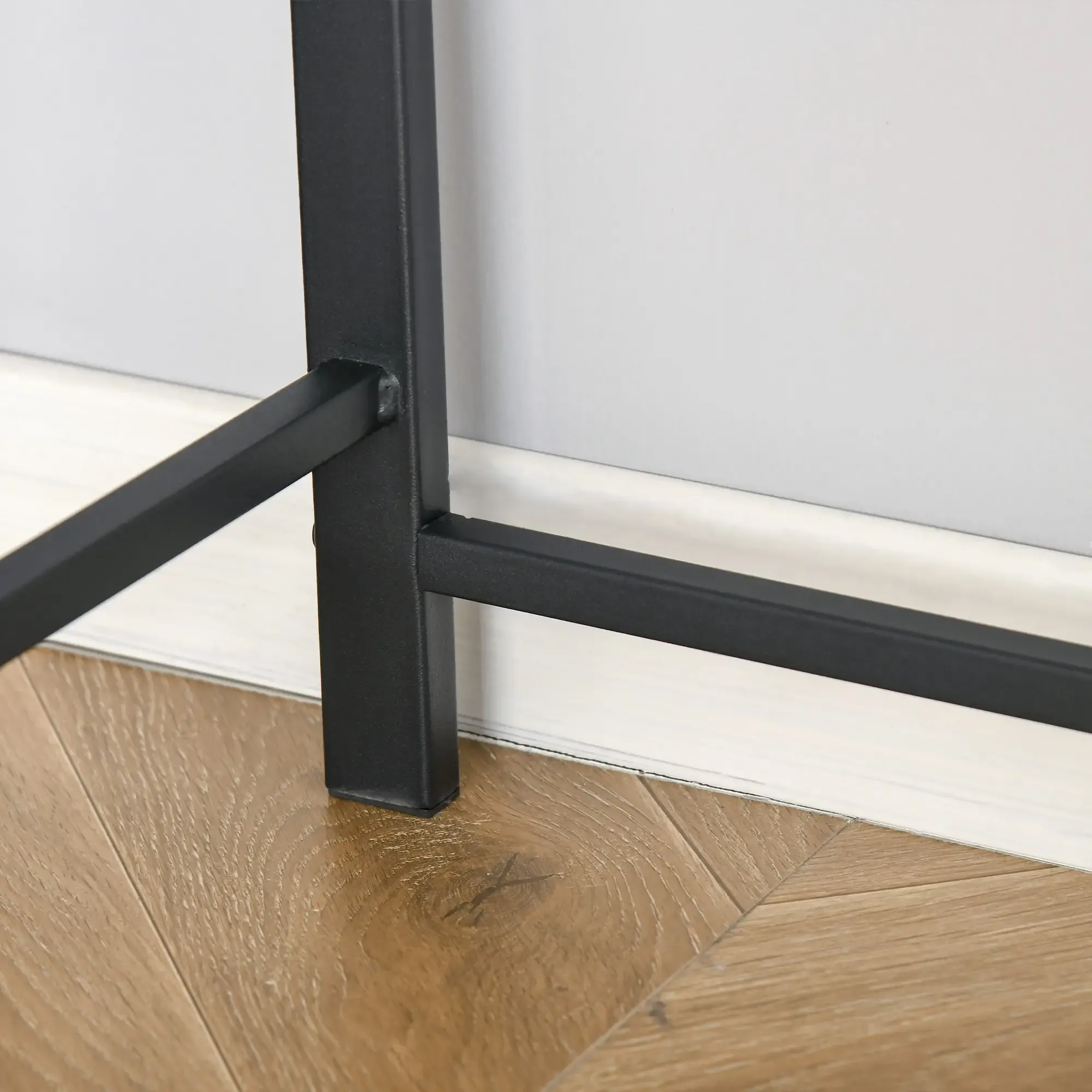 Schwarzes Metall-Tischbein auf Holzfußboden. Weiße Wand im Hintergrund. Das Holz hat diagonale Linien.