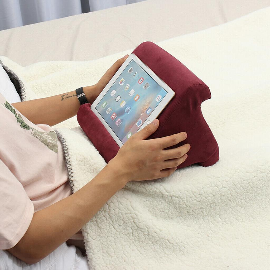 Person hält Tablet in einem weinroten Kissen auf einer weißen Decke. Hände sind sichtbar, trägt eine Uhr. Tablet-Bildschirm ist an.