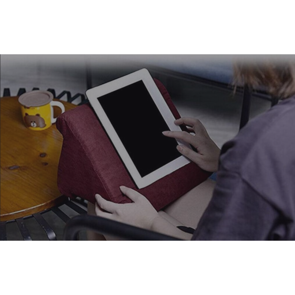 Eine Person benutzt ein Tablet auf einem roten Kissen. Ein gelber Becher mit einem Bären steht auf einem Tisch.