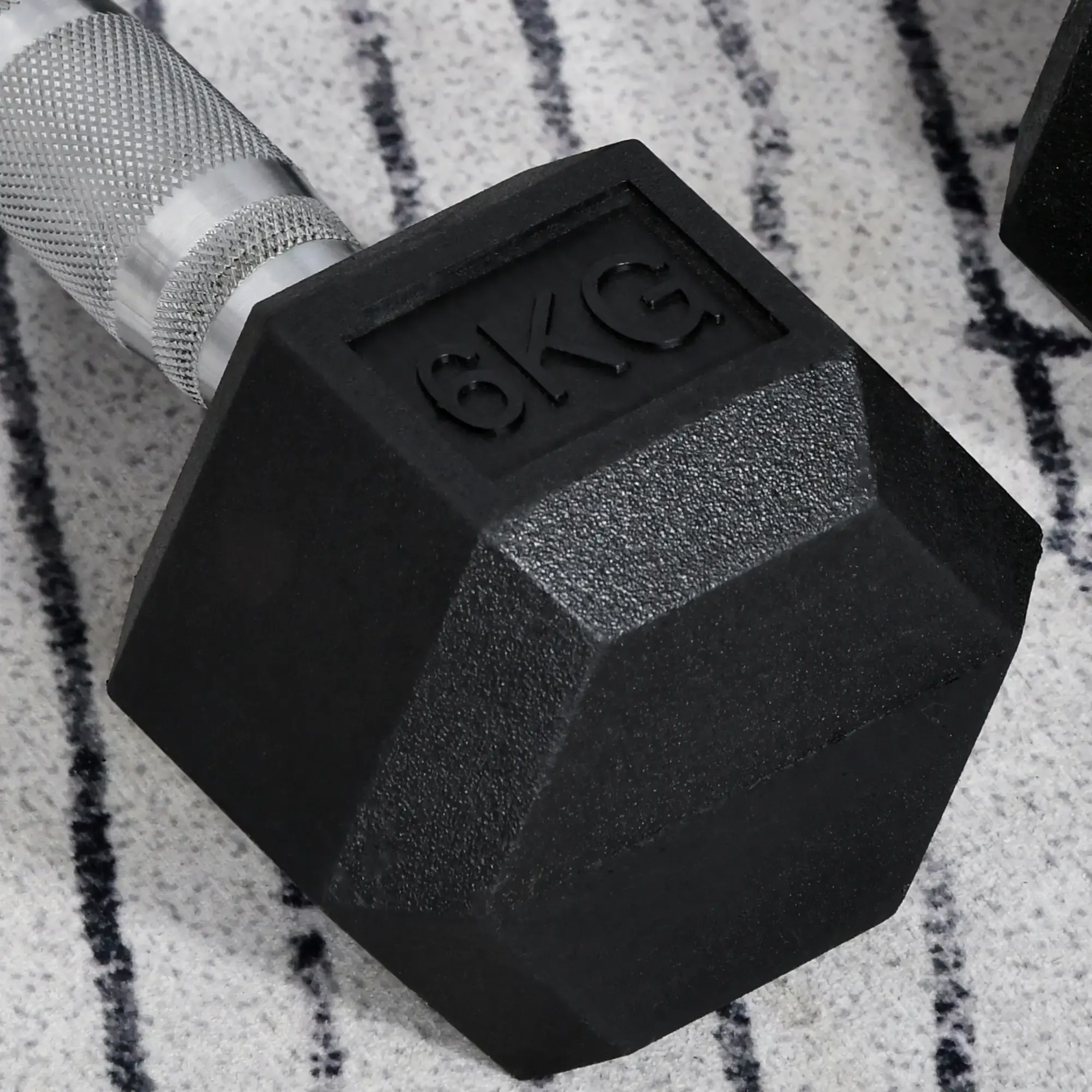 Eine schwarze 6KG-Hantel mit silbernem Griff liegt auf einem gemusterten grauen Teppich. Die Hantel ist sechseckig.