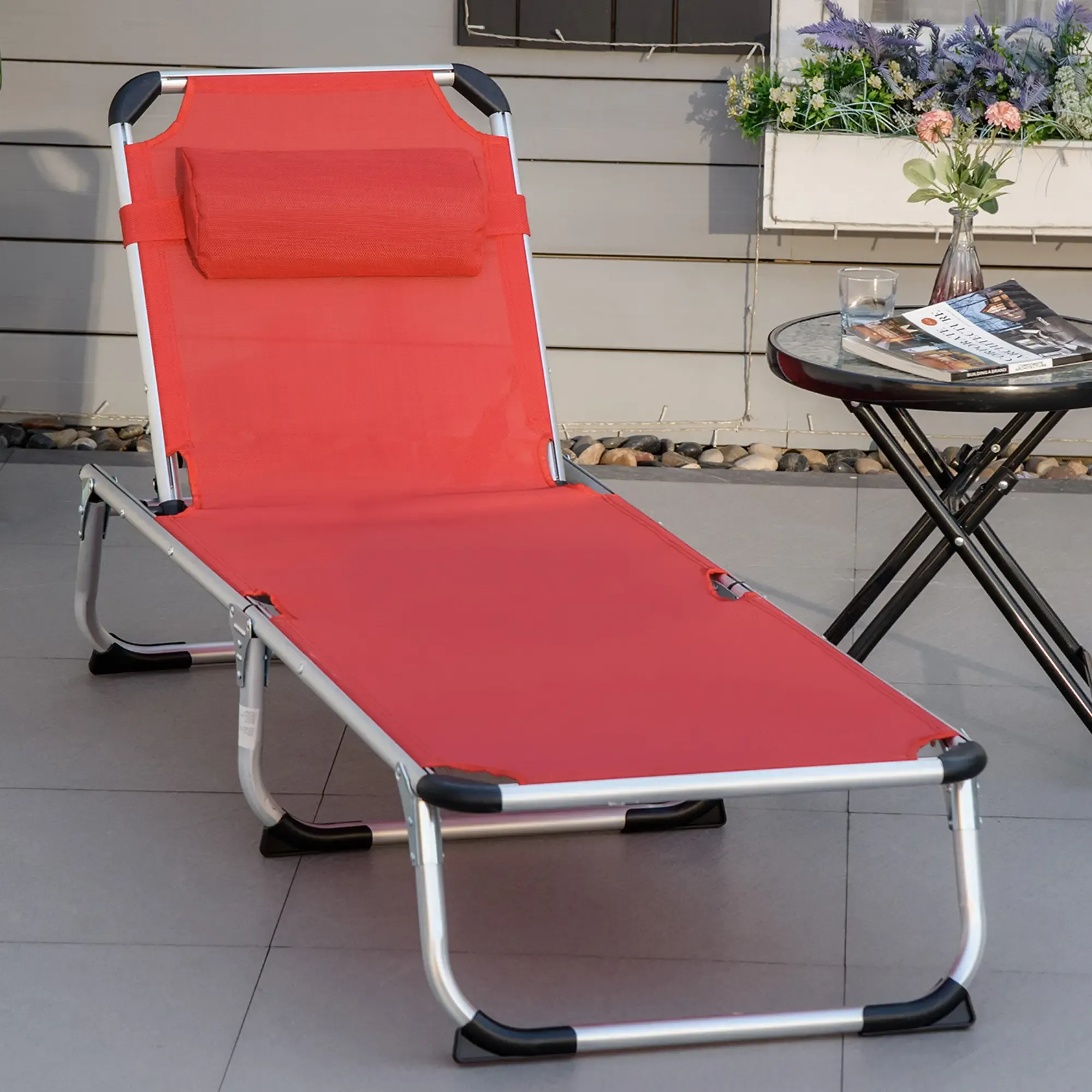 Ein roter Klappliegestuhl steht auf einer Terrasse. Im Hintergrund stehen ein kleiner Tisch und Blumen.
