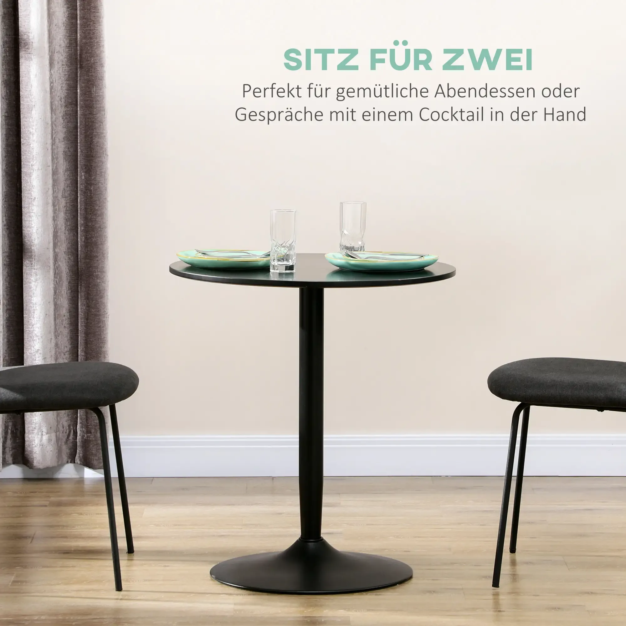 Ein kleiner, runder, schwarzer Tisch ist für zwei Personen mit Tellern, Gläsern und Stühlen gedeckt. Der Hintergrund sind helle Wände und Vorhänge.