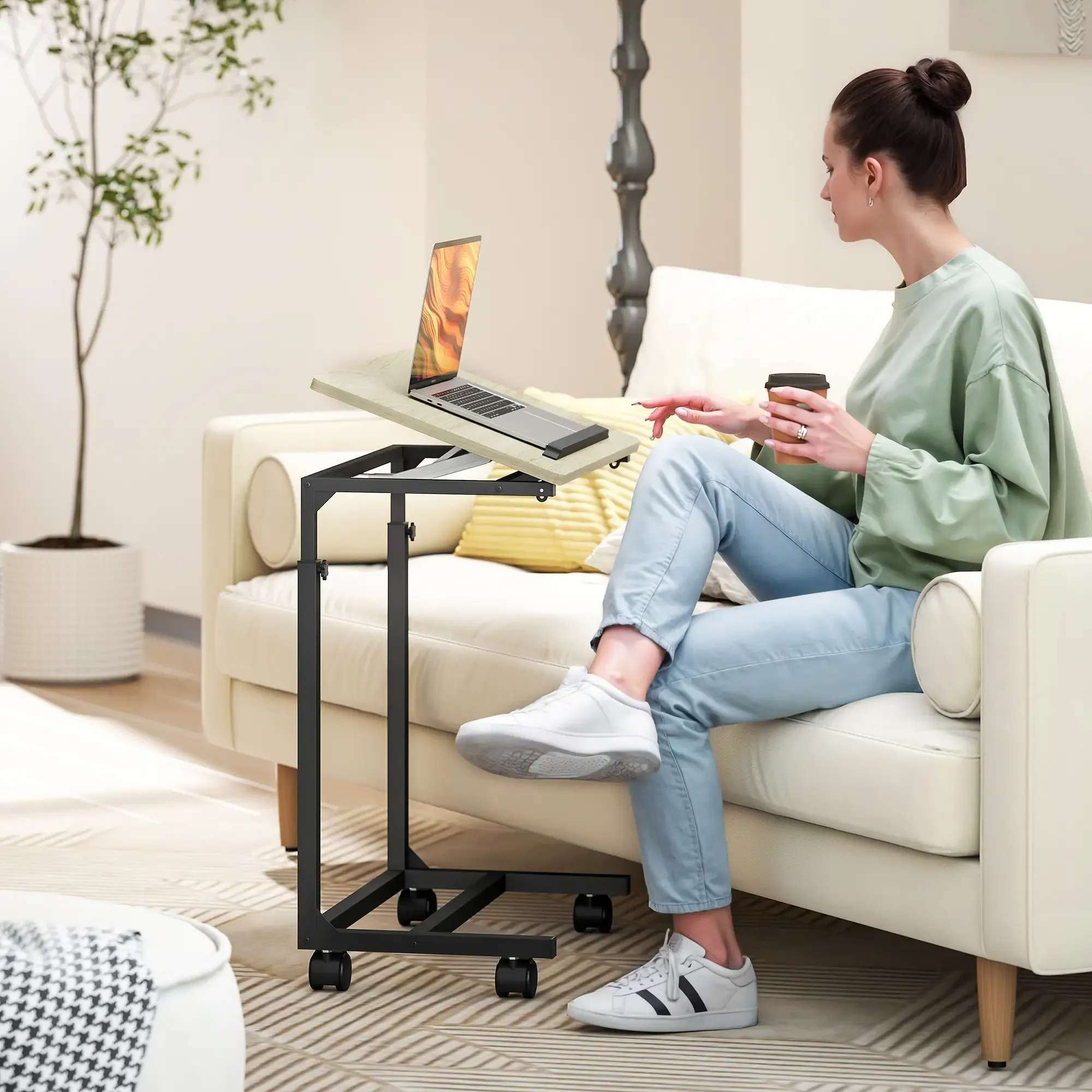 Eine Frau sitzt auf einem Sofa mit einem Laptop auf einem Tisch. Sie hält eine Kaffeetasse. Eine Pflanze und eine Lampe sind im Hintergrund.
