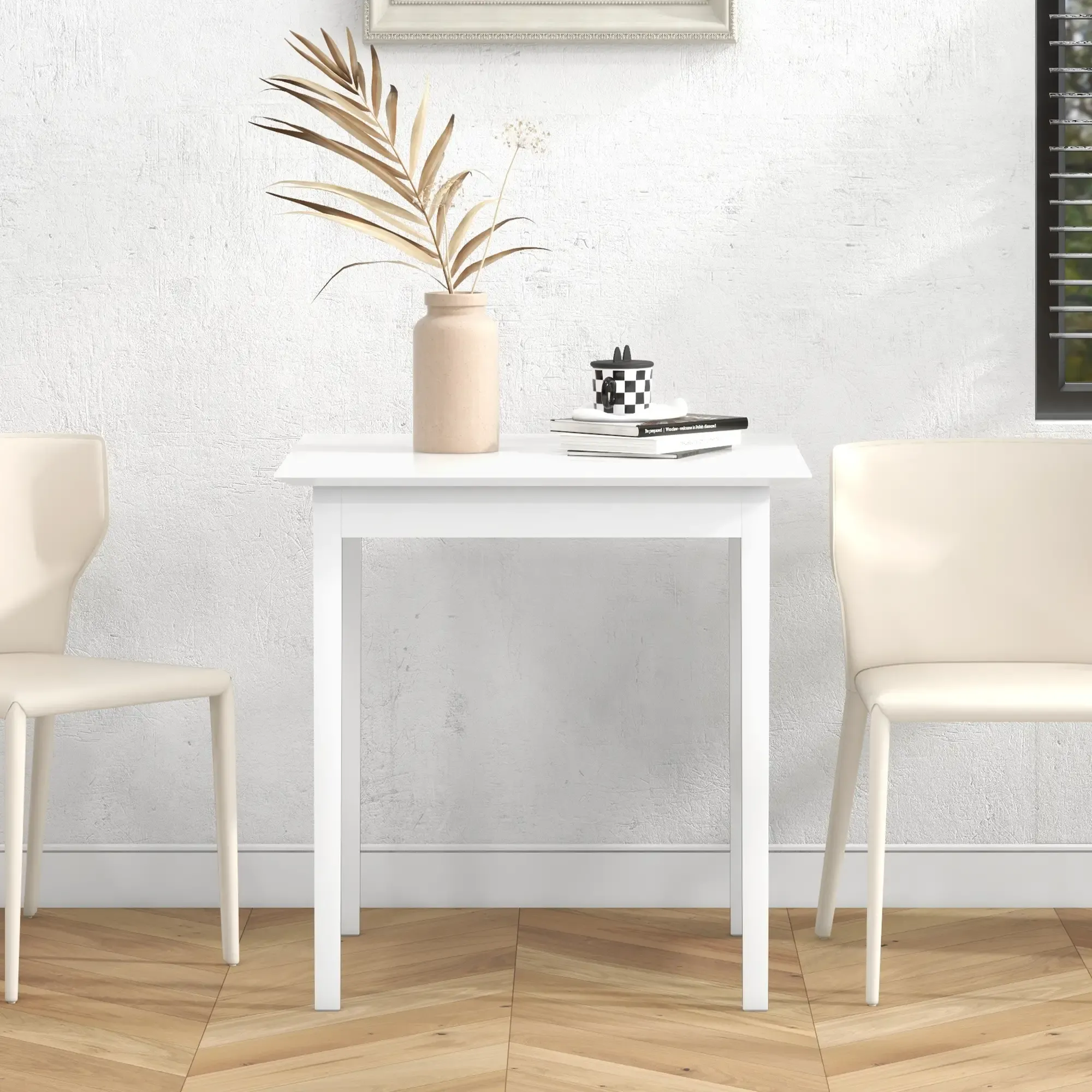 Ein weißer quadratischer Tisch mit zwei Stühlen. Auf dem Tisch eine Vase mit getrockneten Blättern und eine karierte Tasse. Hintergrund: weiße Wand.