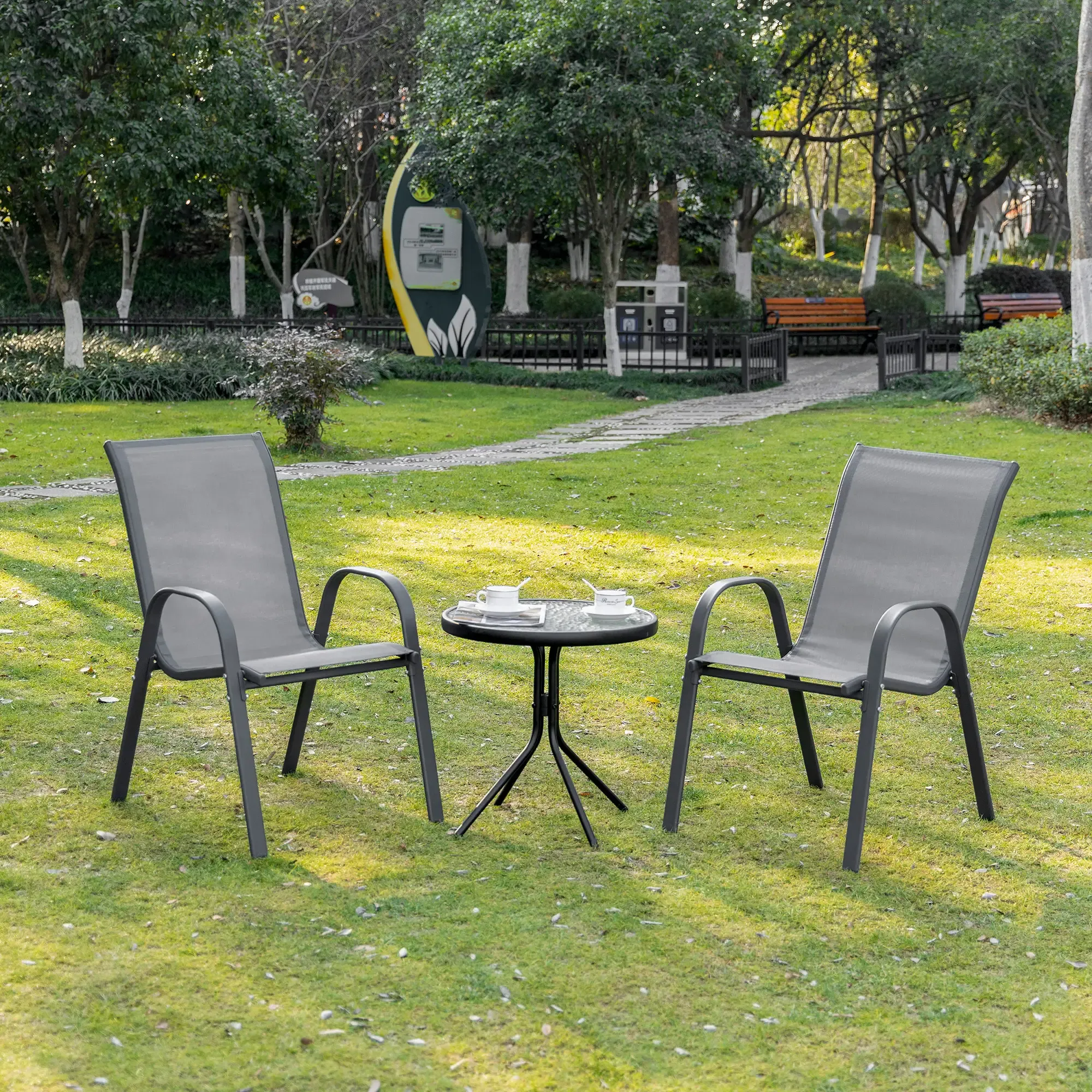 Zwei graue Stühle stehen vor einem kleinen Tisch mit Kaffeetassen in einem grasbewachsenen Park. Bäume und Bänke sind im Hintergrund.