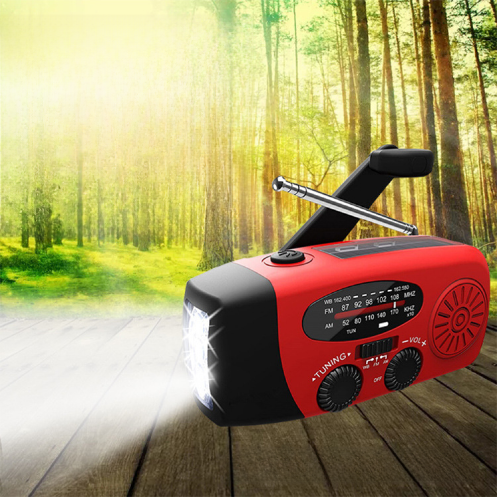 Ein rotes und schwarzes Notfallradio mit einer Taschenlampe steht auf einer Holzoberfläche, der Hintergrund ist ein grüner Wald mit Sonnenlicht.