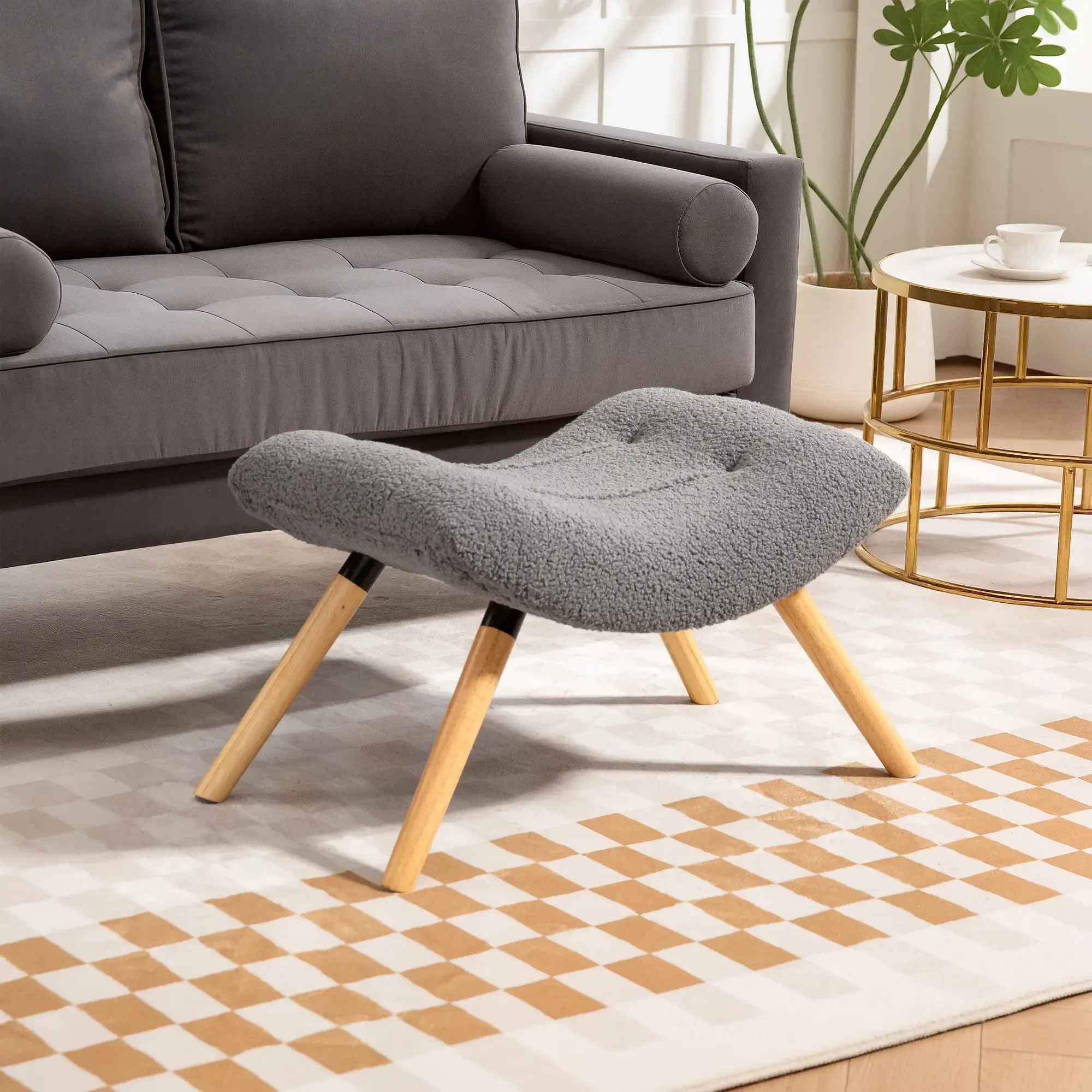 Ein grauer Fußhocker mit Holzbeinen steht auf einem karierten Teppich. Im Hintergrund sind ein graues Sofa, ein Beistelltisch und eine Pflanze zu sehen.