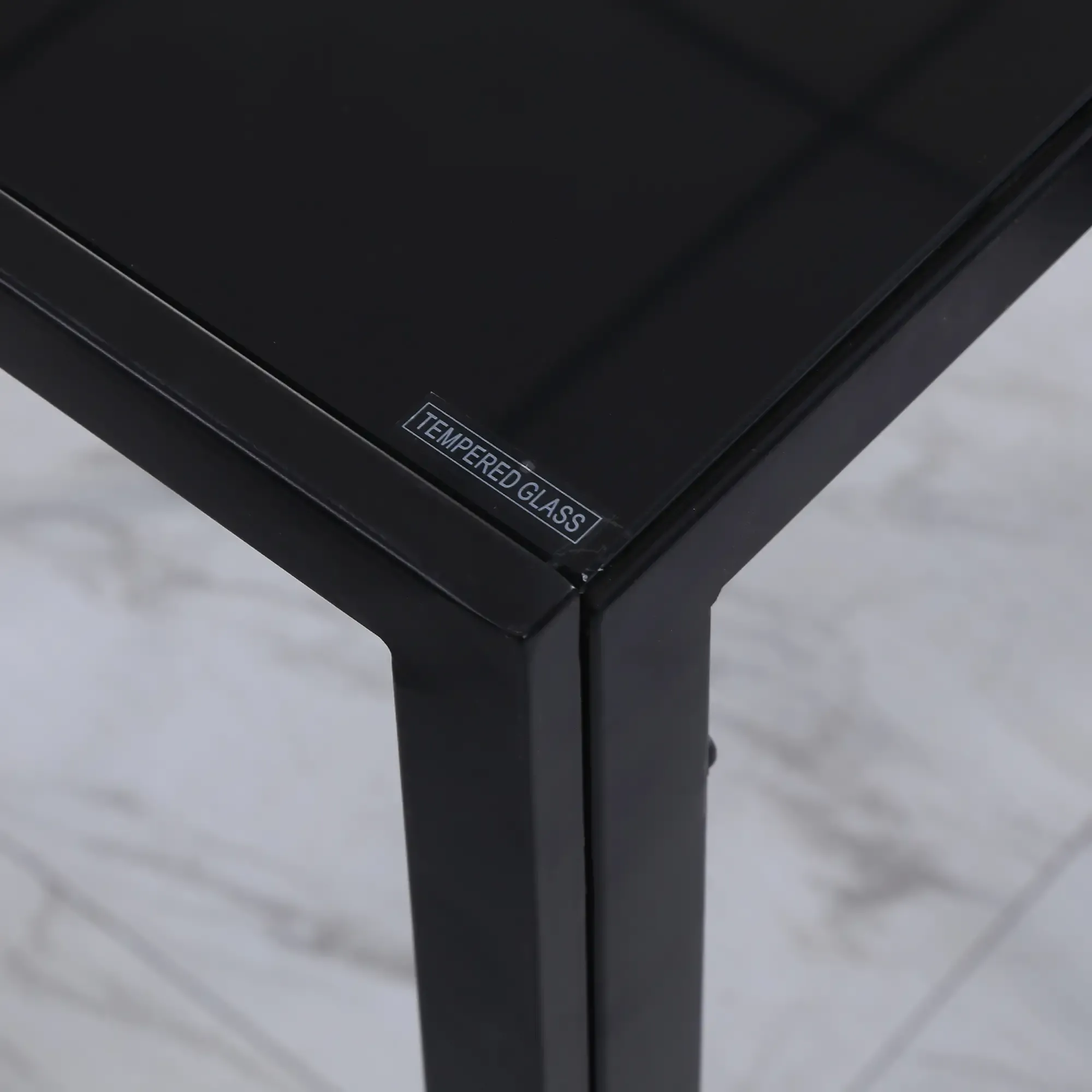 Nahaufnahme einer schwarzen Tisch-Ecke. Sie hat eine Glasplatte mit "TEMPERED GLASS"-Etikett. Die Beine sind ebenfalls schwarz.