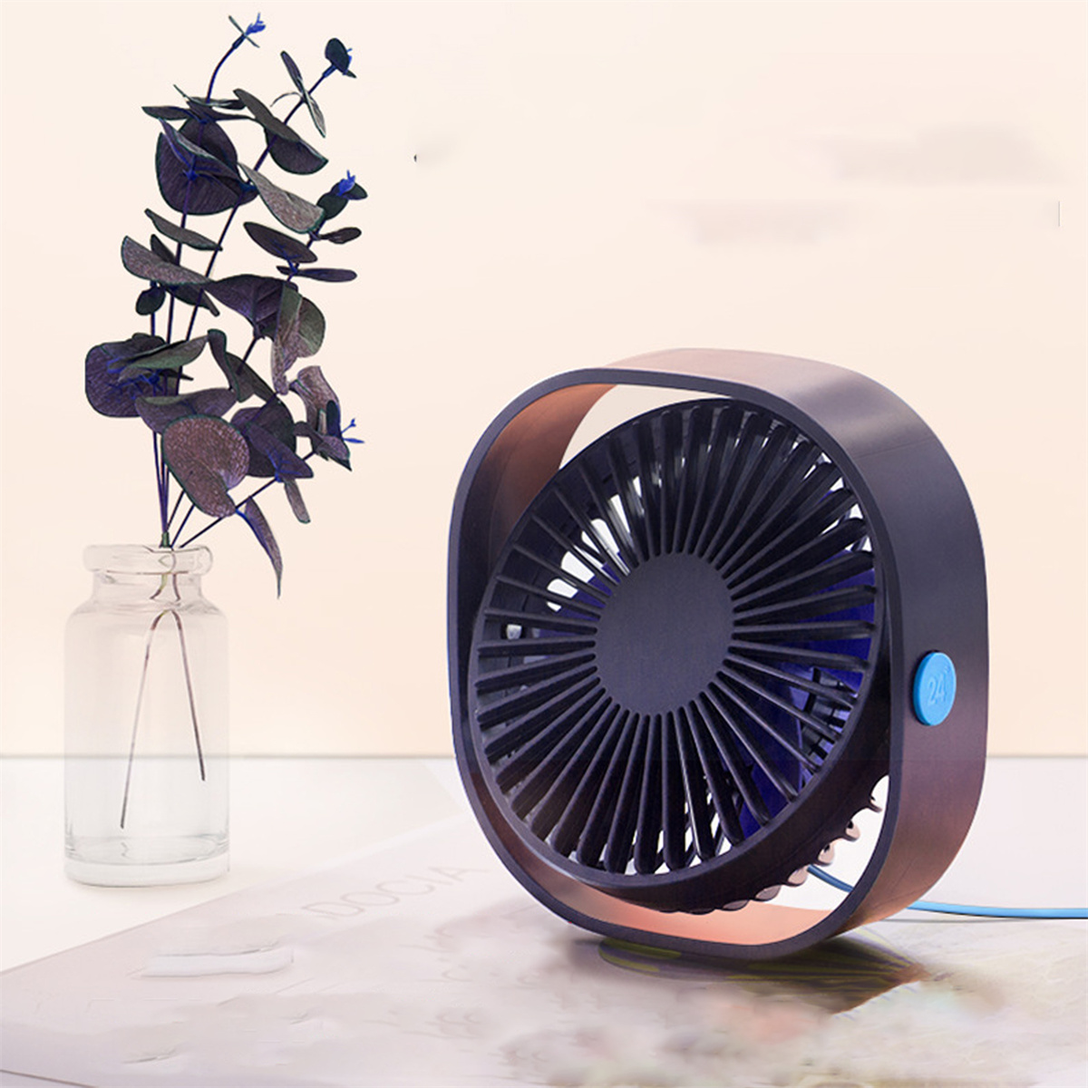 Ein kleiner, dunkelblauer Ventilator mit blauem Knopf. Eine Glasvase enthält lila Blätter. Der Hintergrund ist hellpfirsichfarben.