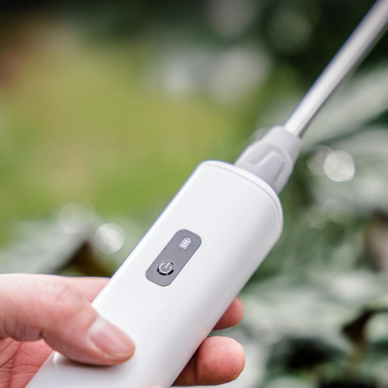 Eine Hand hält ein weißes Gerät mit einem silbernen Stab. Grünes Laub ist im Hintergrund unscharf. Das Gerät hat Strom- und Batteriesymbole.