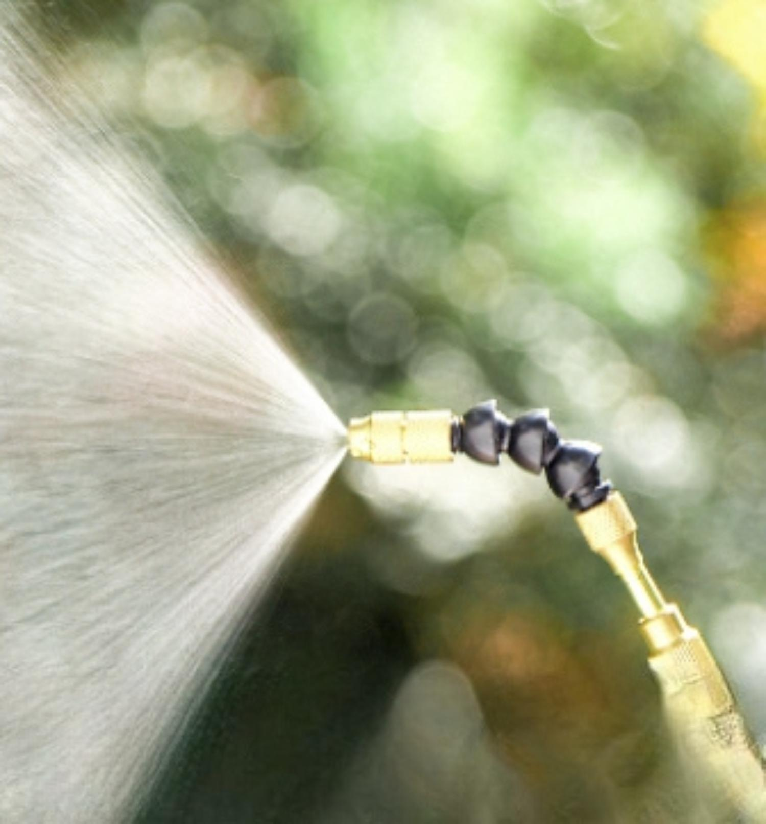 Eine Messingdüse sprüht Wasser, mit einem schwarzen, flexiblen Gelenk, vor einem verschwommenen grünen Hintergrund aus Laub.