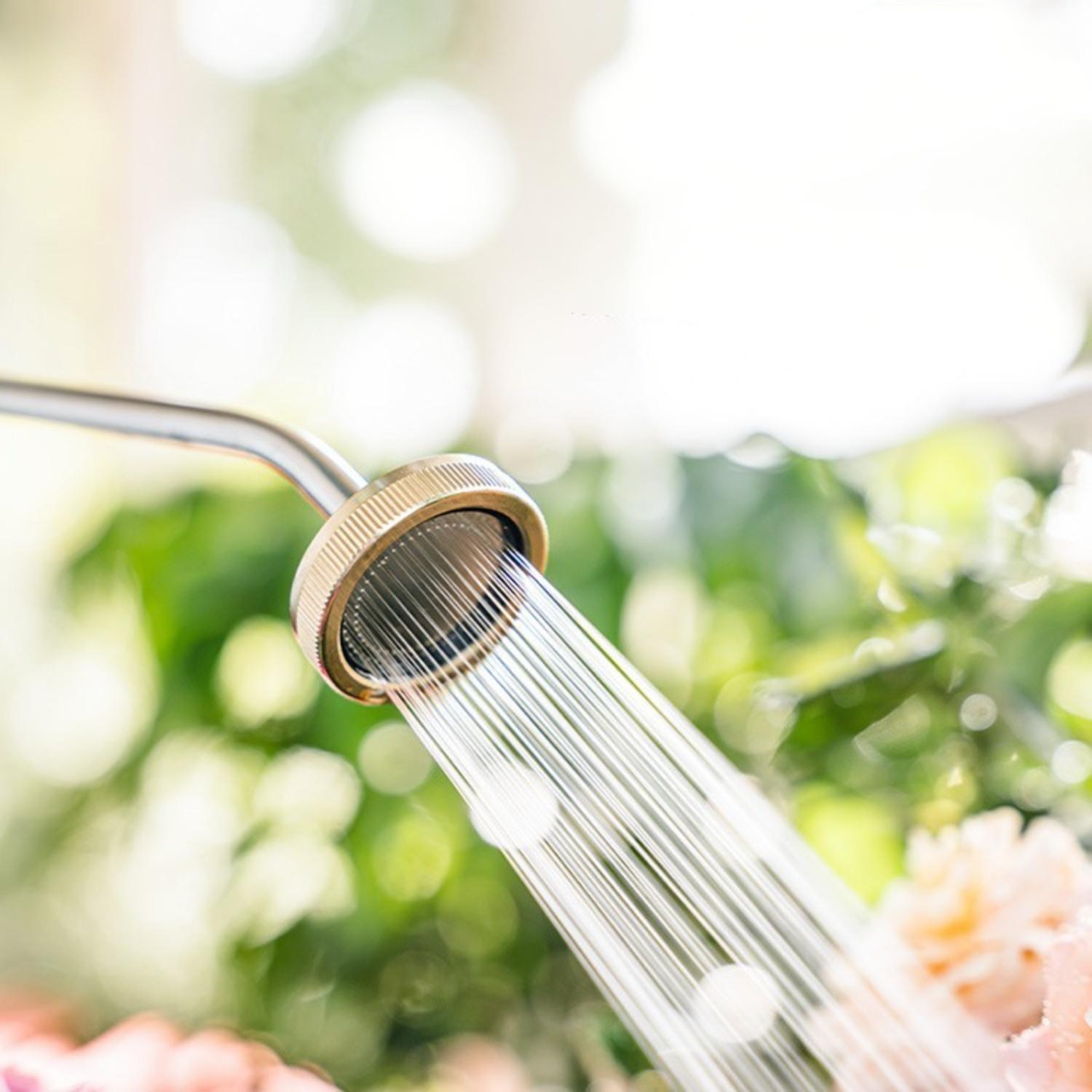 Ein Duschkopf sprüht Wasser. Grünes Laub im Hintergrund. Einige rosa Blumen im Vordergrund. Der Duschkopf ist metallisch.