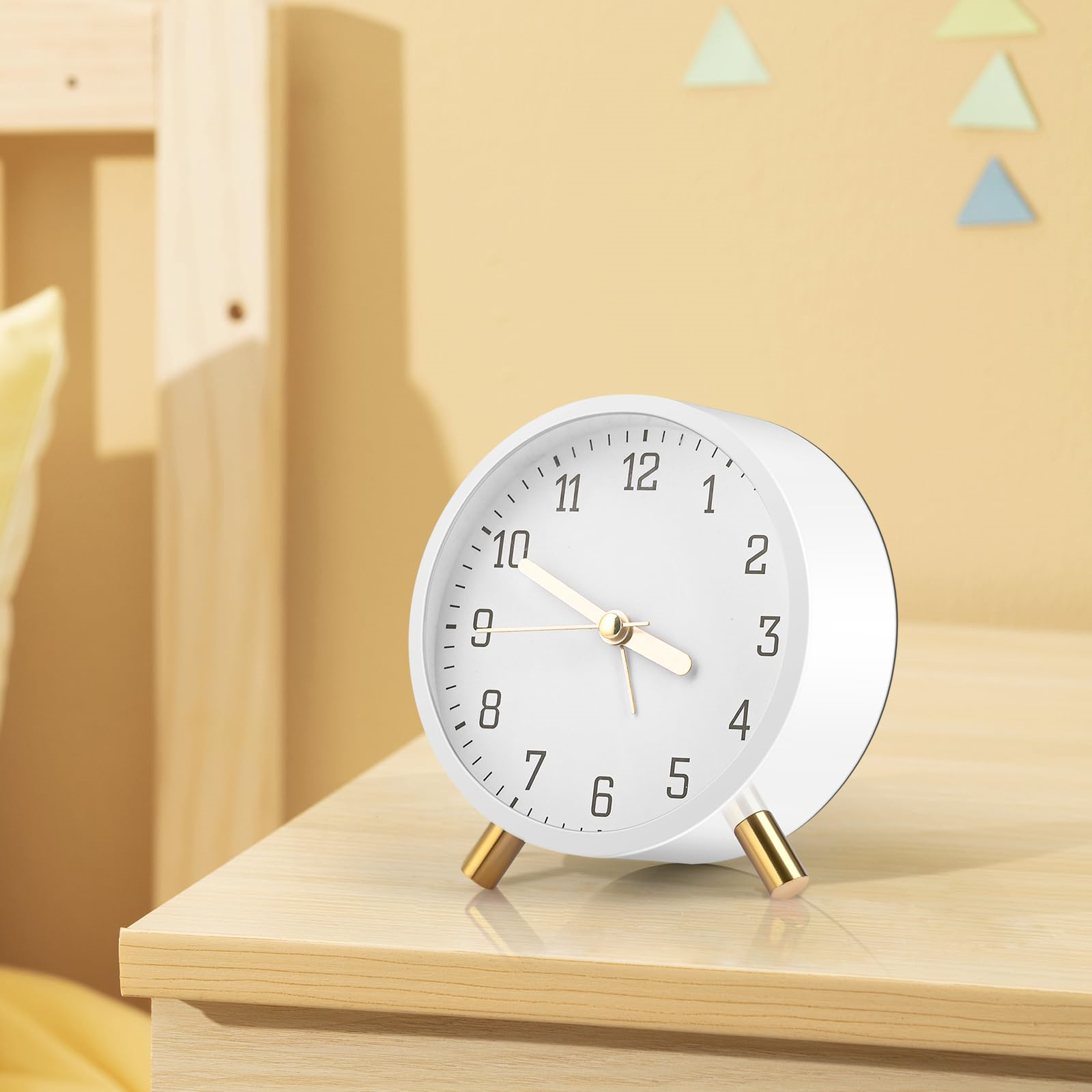 Ein weißer Wecker mit goldenen Beinen auf einer Holzoberfläche. Die Uhr hat schwarze Zahlen und Zeiger. Dreiecke an der Wand.