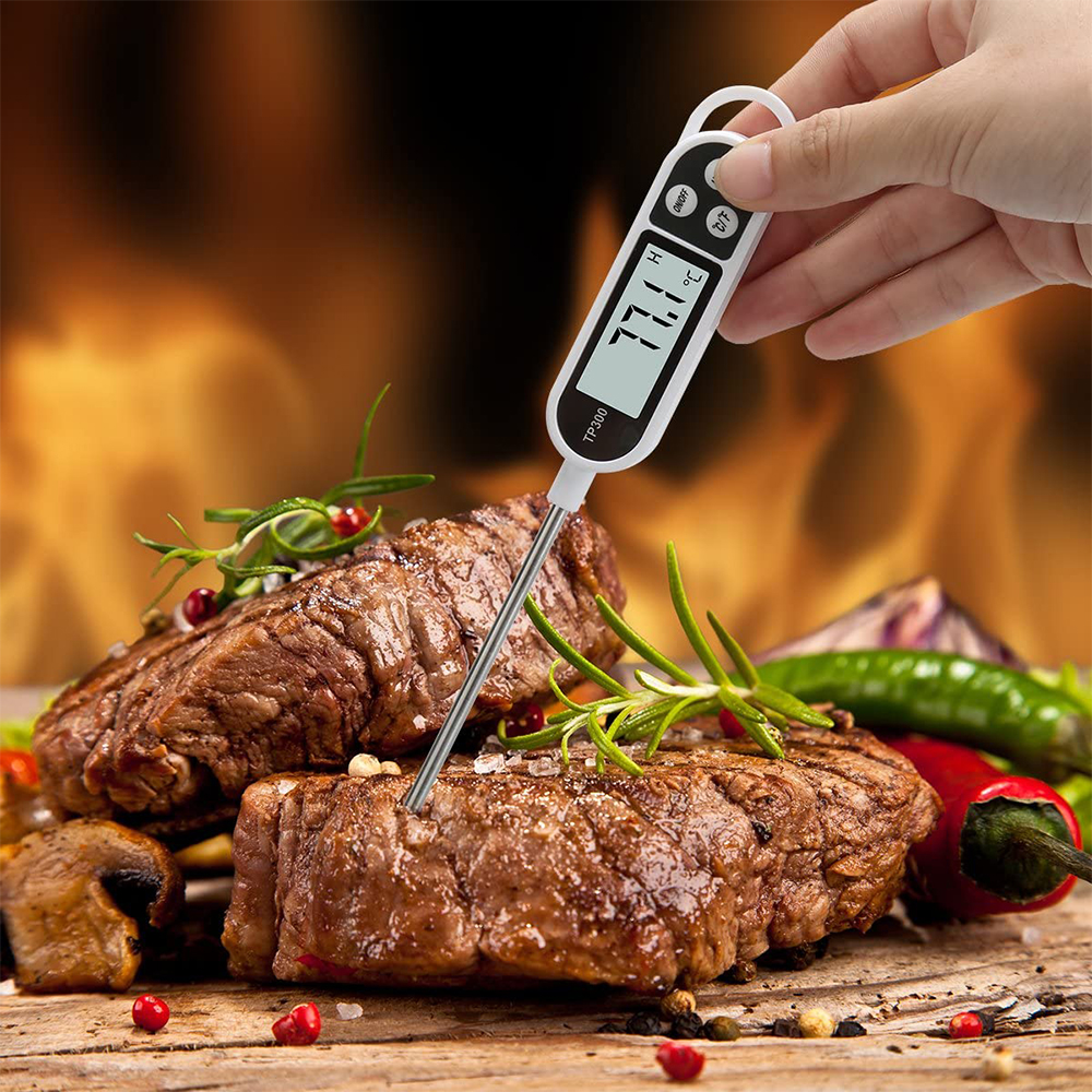 Eine Hand hält ein Fleischthermometer in einem Steak. Das Thermometer zeigt 77,1 Grad an. Rosmarin und Paprika sind vorhanden.
