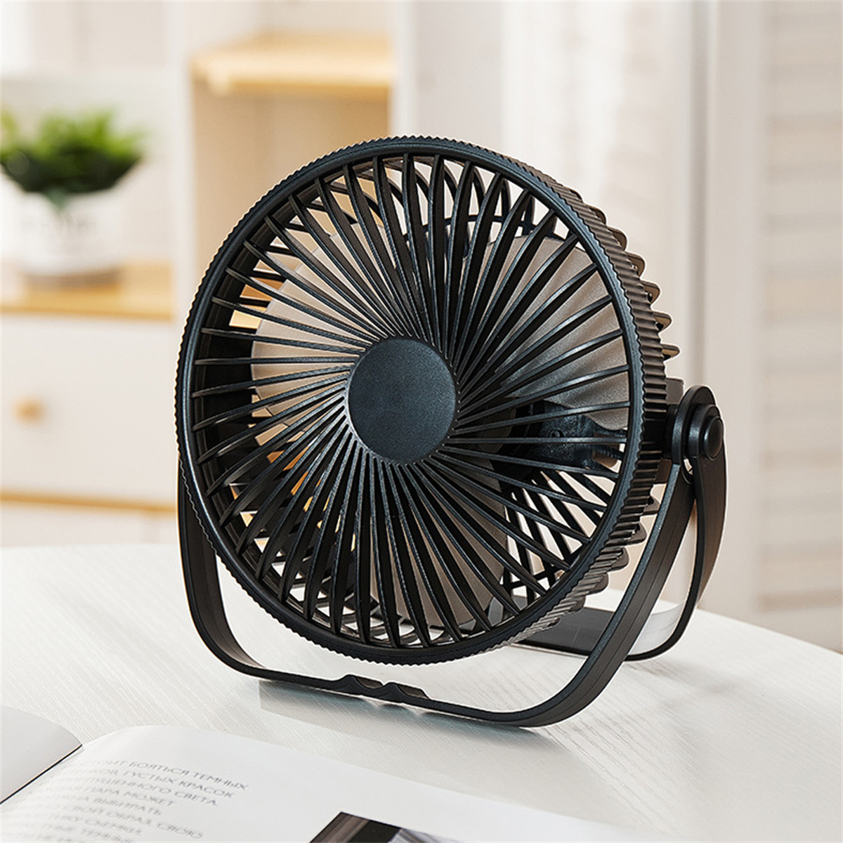 Ein schwarzer Tischventilator steht auf einem weißen Tisch. Der Ventilator ist eingeschaltet, die Flügel sind sichtbar. Im Hintergrund ist eine Pflanze.