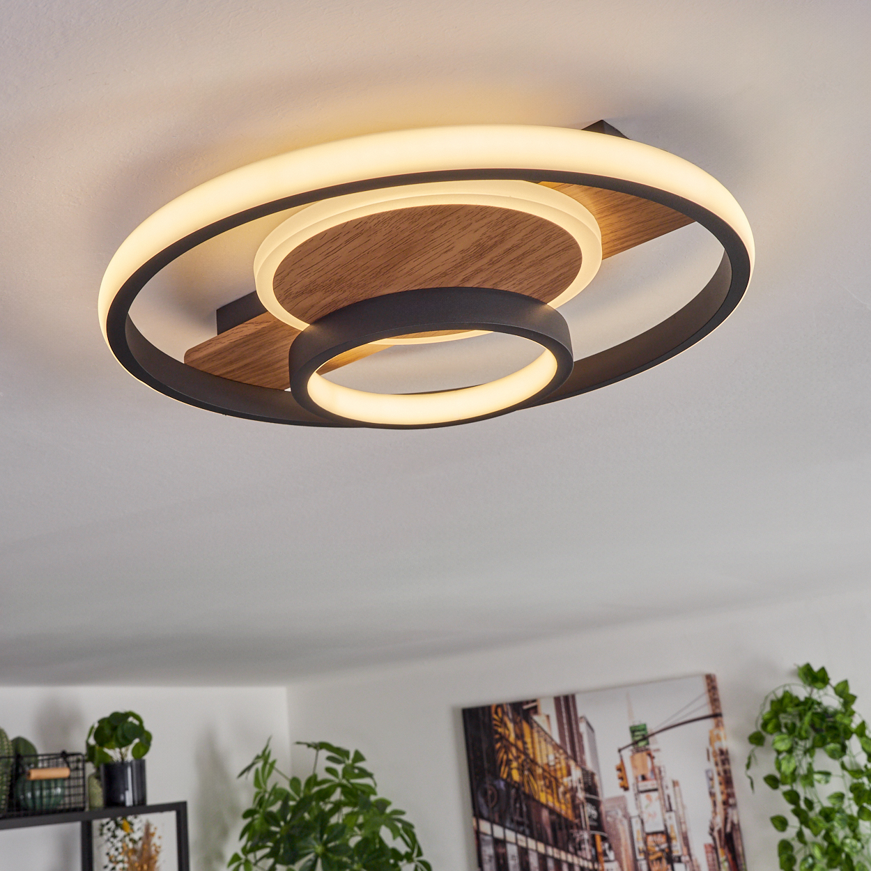 Moderne Deckenleuchte mit schwarzen Ringen und Holzdetails. Warmes Licht beleuchtet den Raum. Pflanzen und Kunstwerke darunter sichtbar.