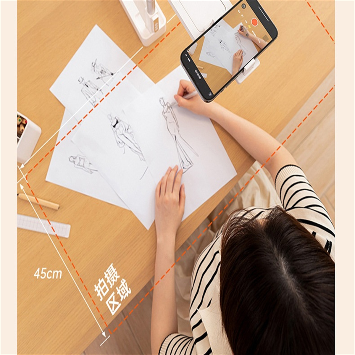 Eine Person skizziert Modedesigns auf Papier, gefilmt von einem Telefon. Zeichnungen, Bleistifte und Lineal liegen auf dem Holztisch. Eine Aufnahme läuft.
