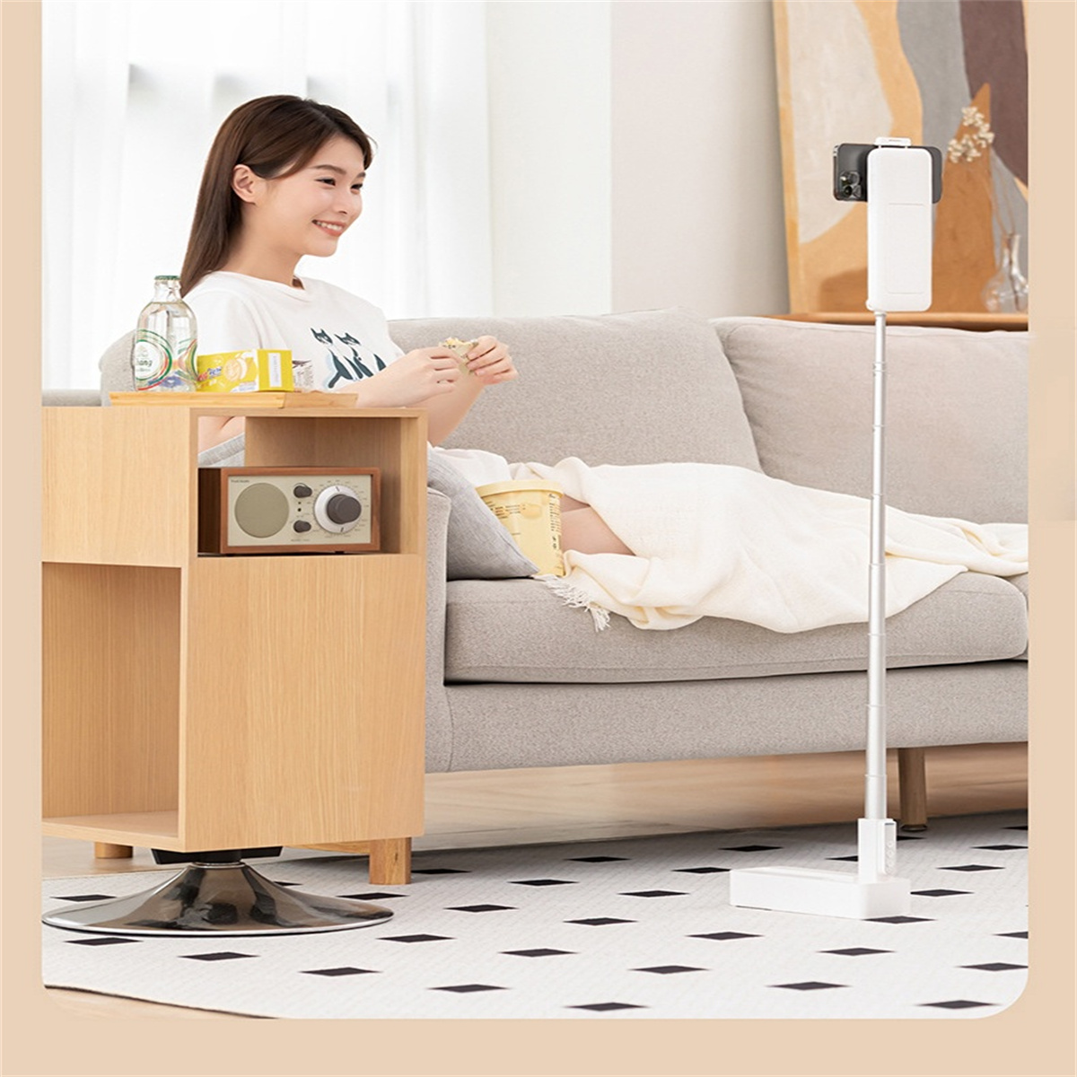 Eine Frau sitzt mit Snacks auf einem Sofa. Ein Telefon auf einem Ständer ist vor ihr. Links ein Tisch mit Radio.