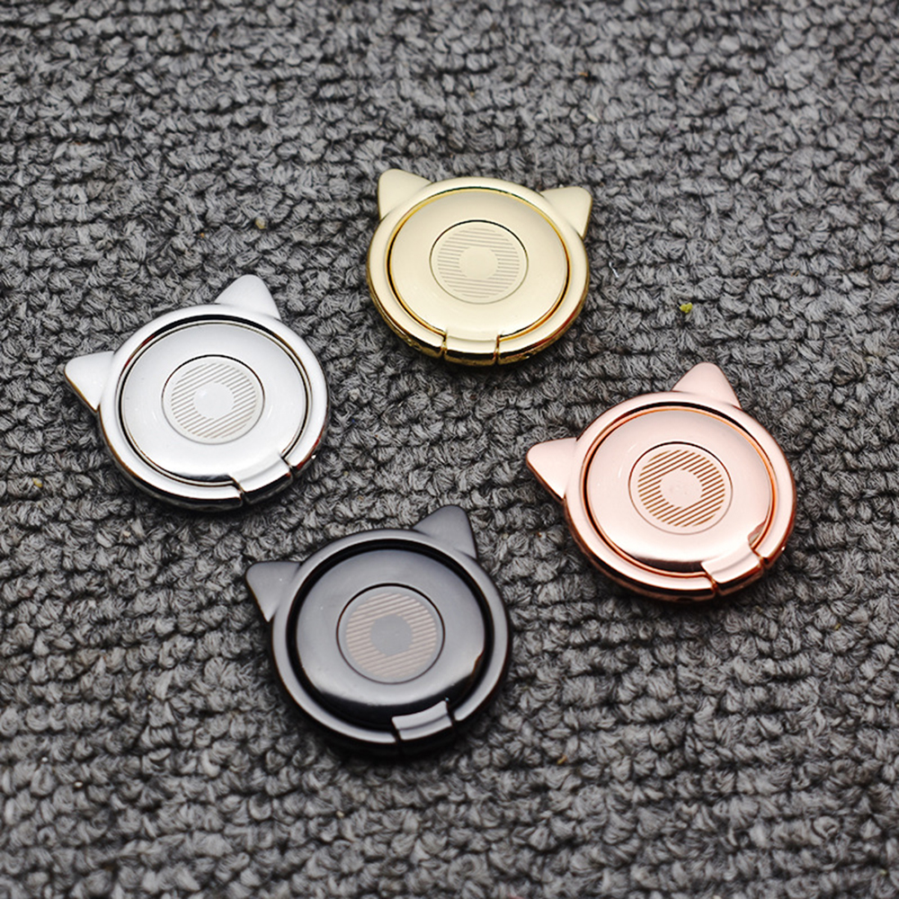 Vier katzenförmige Handyhalter in Silber, Gold, Roségold und Schwarz. Sie liegen auf einer grauen, strukturierten Oberfläche.
