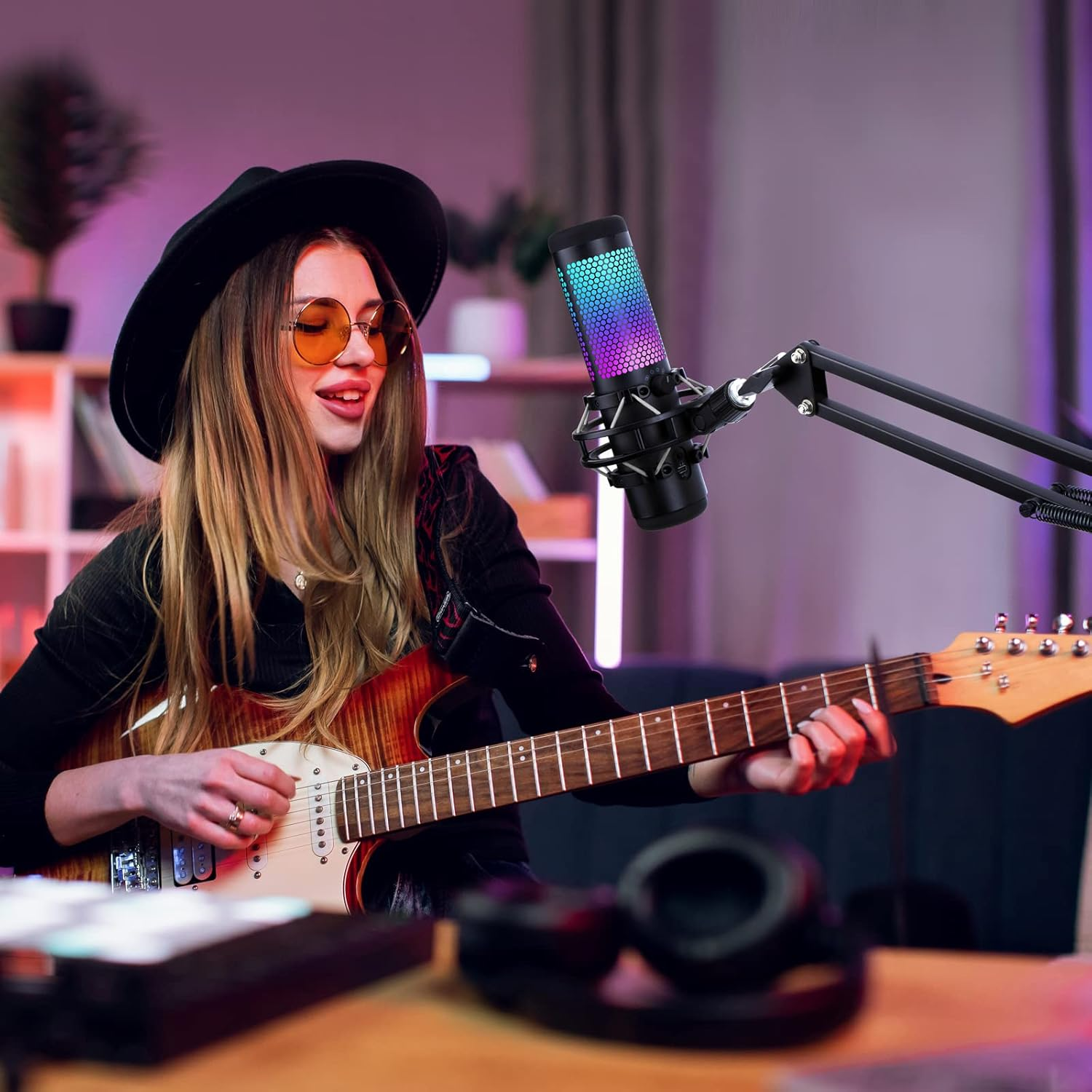 Frau spielt Gitarre mit Mikrofon. Sie trägt Sonnenbrille und Hut. Studio-Setup mit Kopfhörern, Schreibtisch und Bücherregal im Hintergrund.