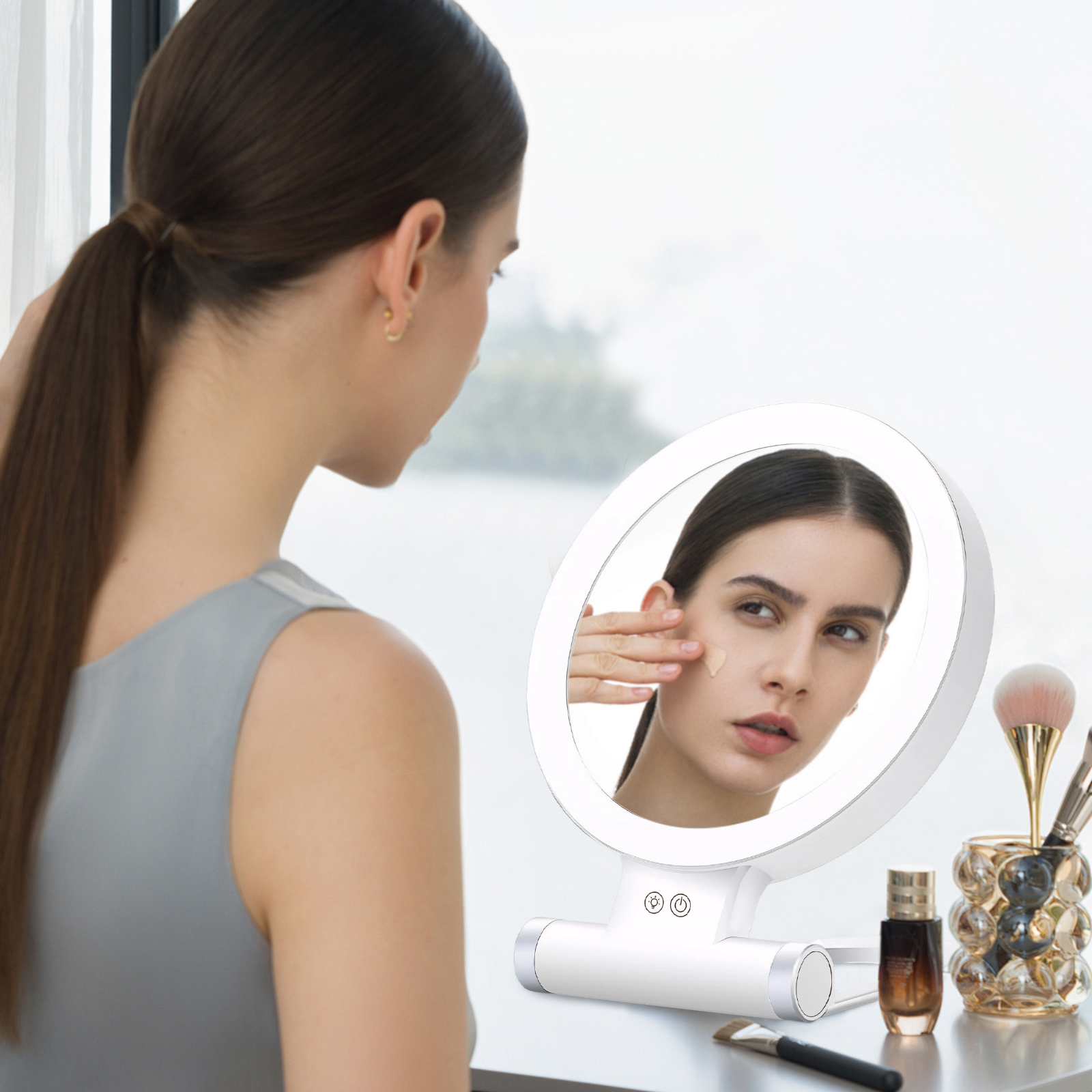 Eine Frau blickt in einen beleuchteten Spiegel und trägt Make-up auf. Pinsel und Schönheitsprodukte stehen auf dem Tisch. Der Hintergrund ist verschwommen.
