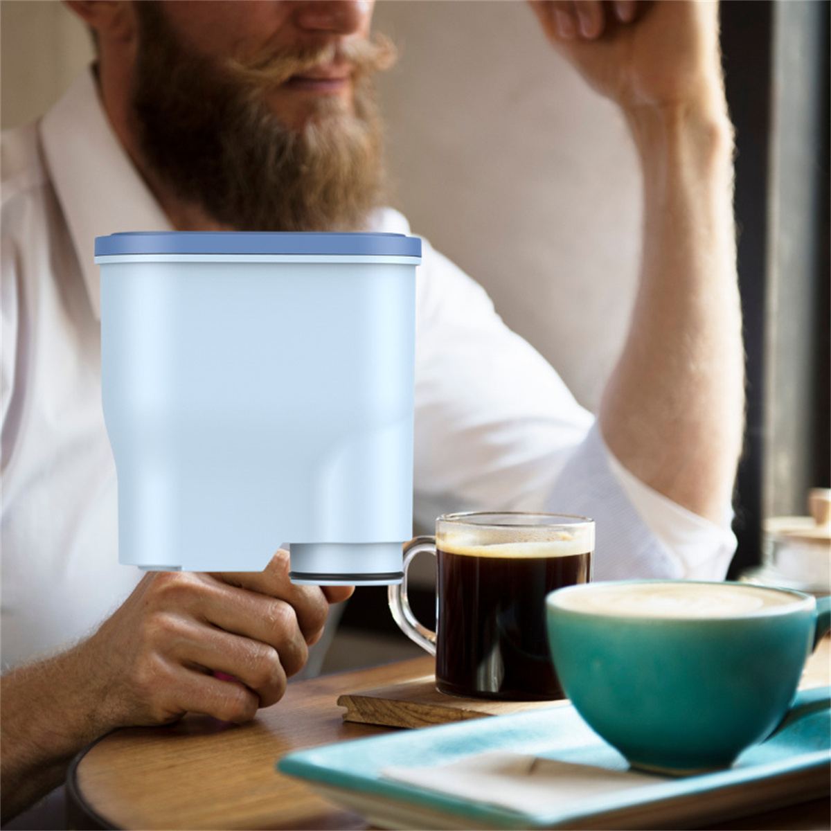 Ein Mann hält einen Wasserfilter. Kaffee in einer Glastasse und eine türkisfarbene Tasse mit Latte stehen auf dem Tisch. Eine Person ist im Hintergrund sichtbar.