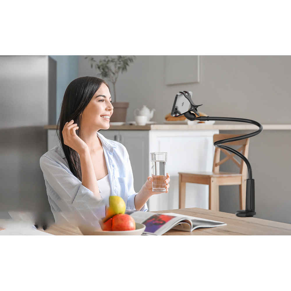 Eine lächelnde Frau hält Wasser, sitzt an einem Tisch mit Obst und einem Buch. Ein Telefon ist an einem flexiblen Arm befestigt.