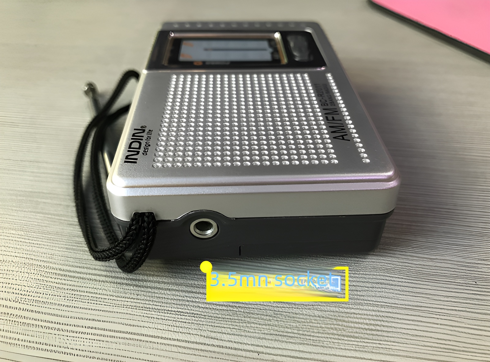 Ein silbernes AM/FM-Radio liegt auf einer grauen Oberfläche. Es hat einen schwarzen Riemen und eine 3,5-mm-Buchse. Im Hintergrund ist eine rosa Fläche.