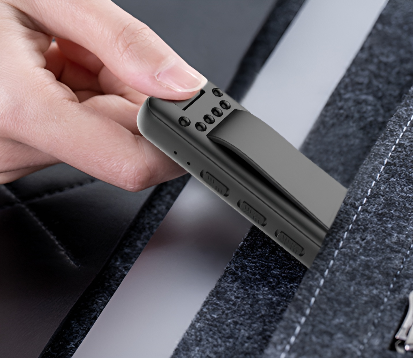Eine Hand steckt ein schwarzes, rechteckiges Gerät mit Clip in eine dunkelgraue Tasche. Das Gerät hat kleine Löcher und Knöpfe.