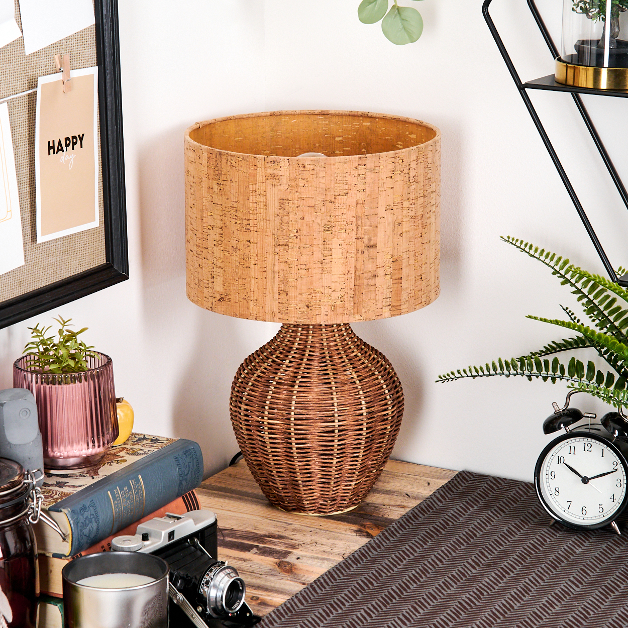 Eine Korblampe auf einem Holztisch mit Büchern, einer Kamera und einer Uhr. Eine 'Happy Day'-Notiz ist im Hintergrund angepinnt.